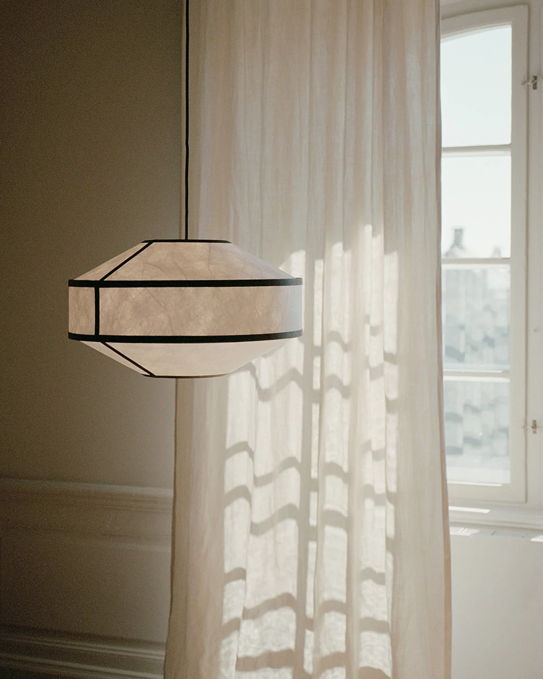 A geometric paper pendant lamp with dark frames hangs beside a sunlit sheer curtain and window.