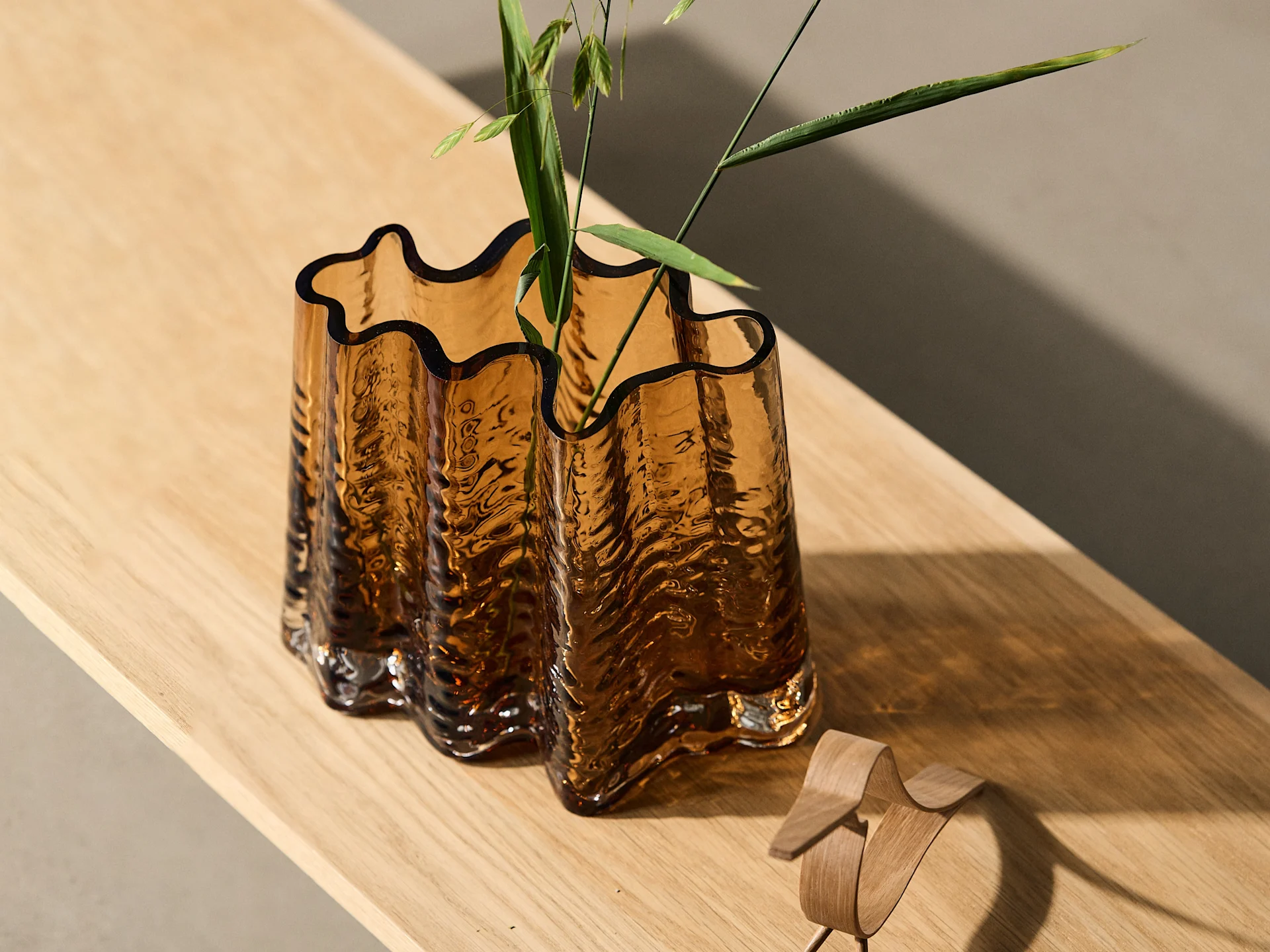 Amber wavy glass vase with green foliage on a light wood surface, next to a small wooden bird.