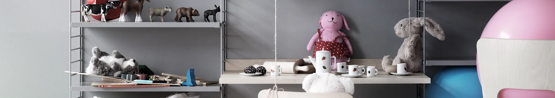 Gray shelves in a child's room hold toys and books. A wooden desk features a pink rabbit doll, a white tea set, and fluffy toy.