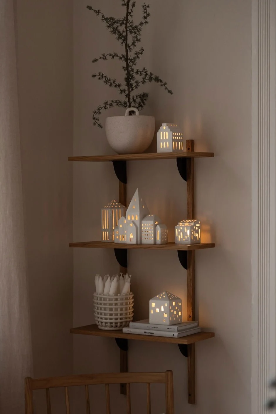 A cozy corner with three wooden shelves displaying white ceramic house lanterns, a vase with branches, and candles in a basket, above a wooden chair with a plaid cushion.