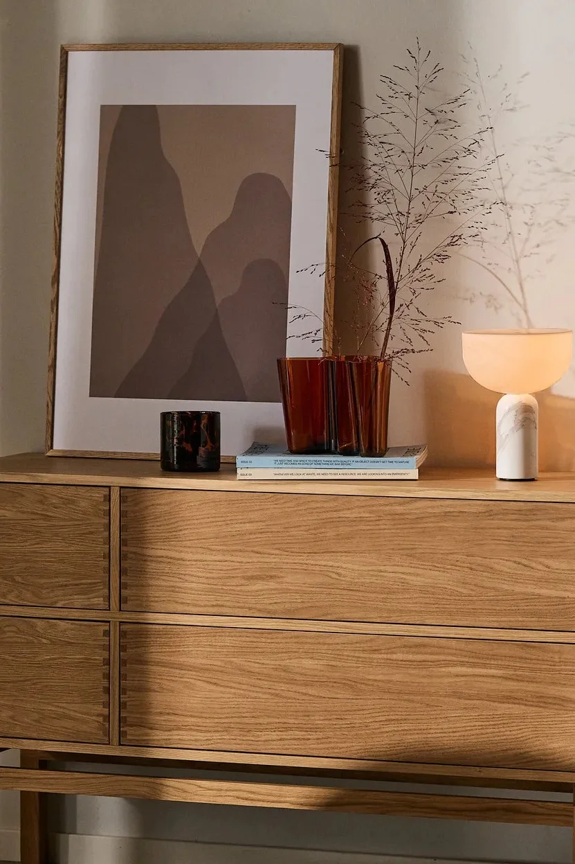 Cozy living space with a fluffy beige armchair, wooden chest of drawers topped with abstract art, vase, and a glowing marble lamp.