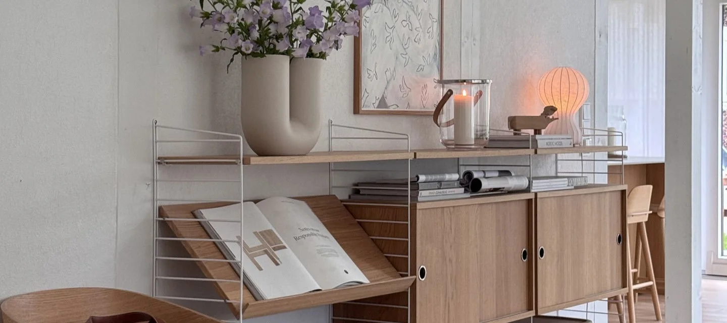 A modular shelving system with white frames and light wood shelves, cabinets, a U-shaped vase with purple flowers, a lit candle in a lantern, and a glowing mushroom-shaped lamp.