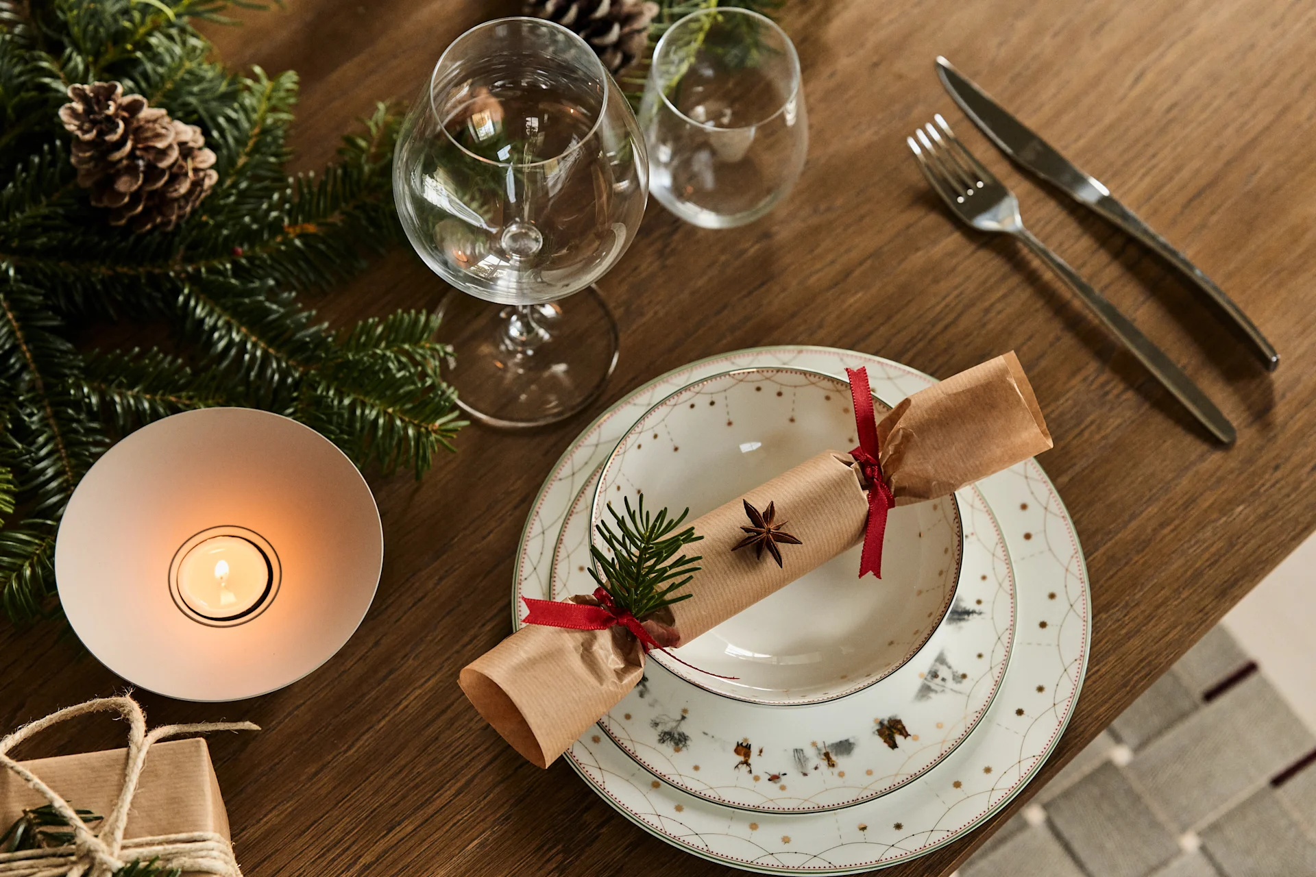 A festive Christmas table setting with a cracker, plates, glasses, cutlery, pine garland, and a lit candle.