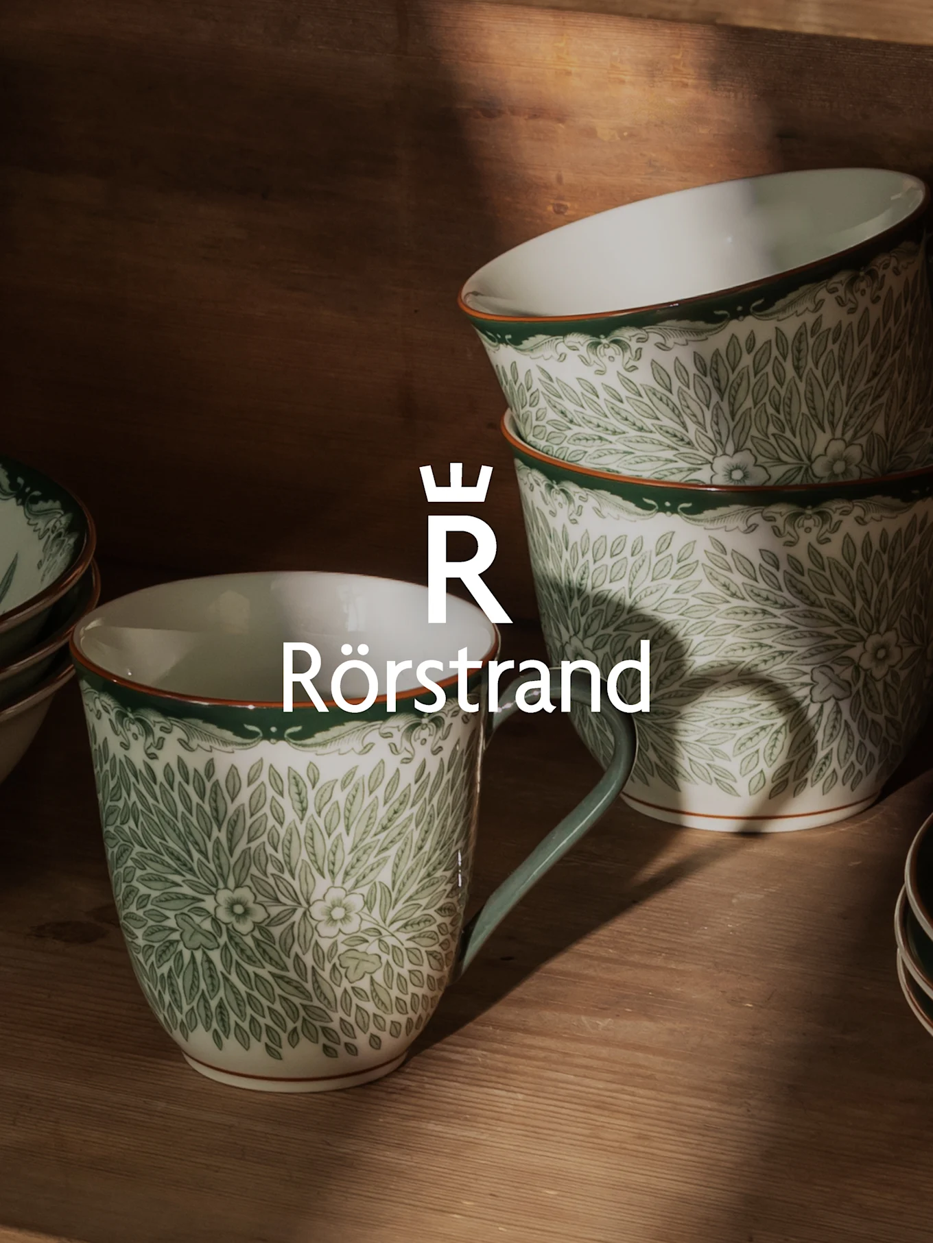 White ceramic mugs and bowls with green leafy pattern, orange rim, and Rörstrand logo.