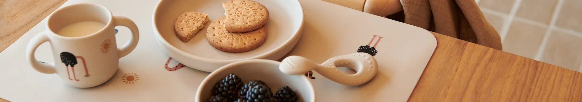 A child's meal with a mug of milk, a bowl of biscuits, a bowl of blackberries, and a baby teether, all on a placemat with ostrich and sun designs on a wooden table.