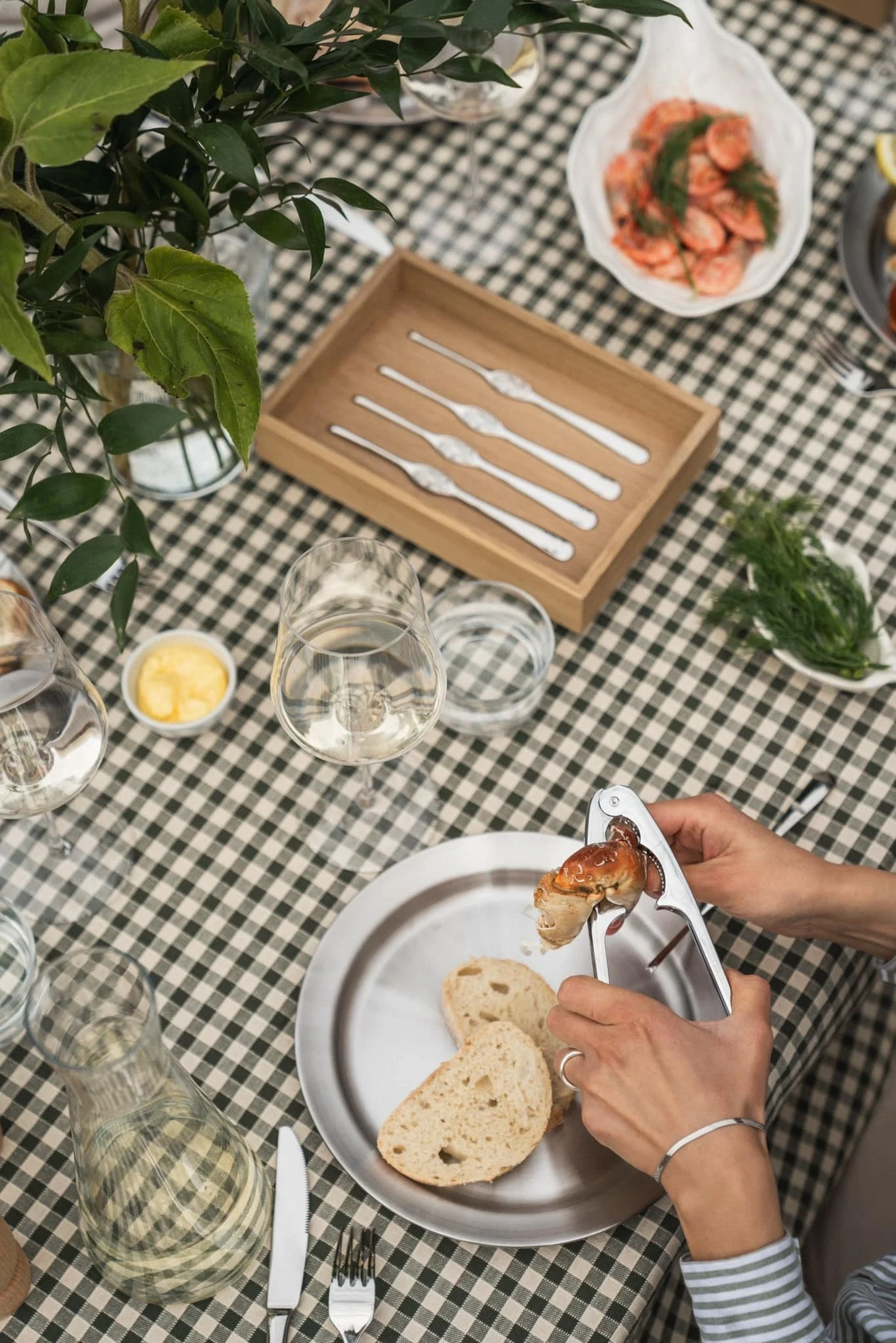 Hands cracking a crab claw with a metal cracker at a table set for a seafood meal, featuring a checkered tablecloth, bread, wine, and shrimp.