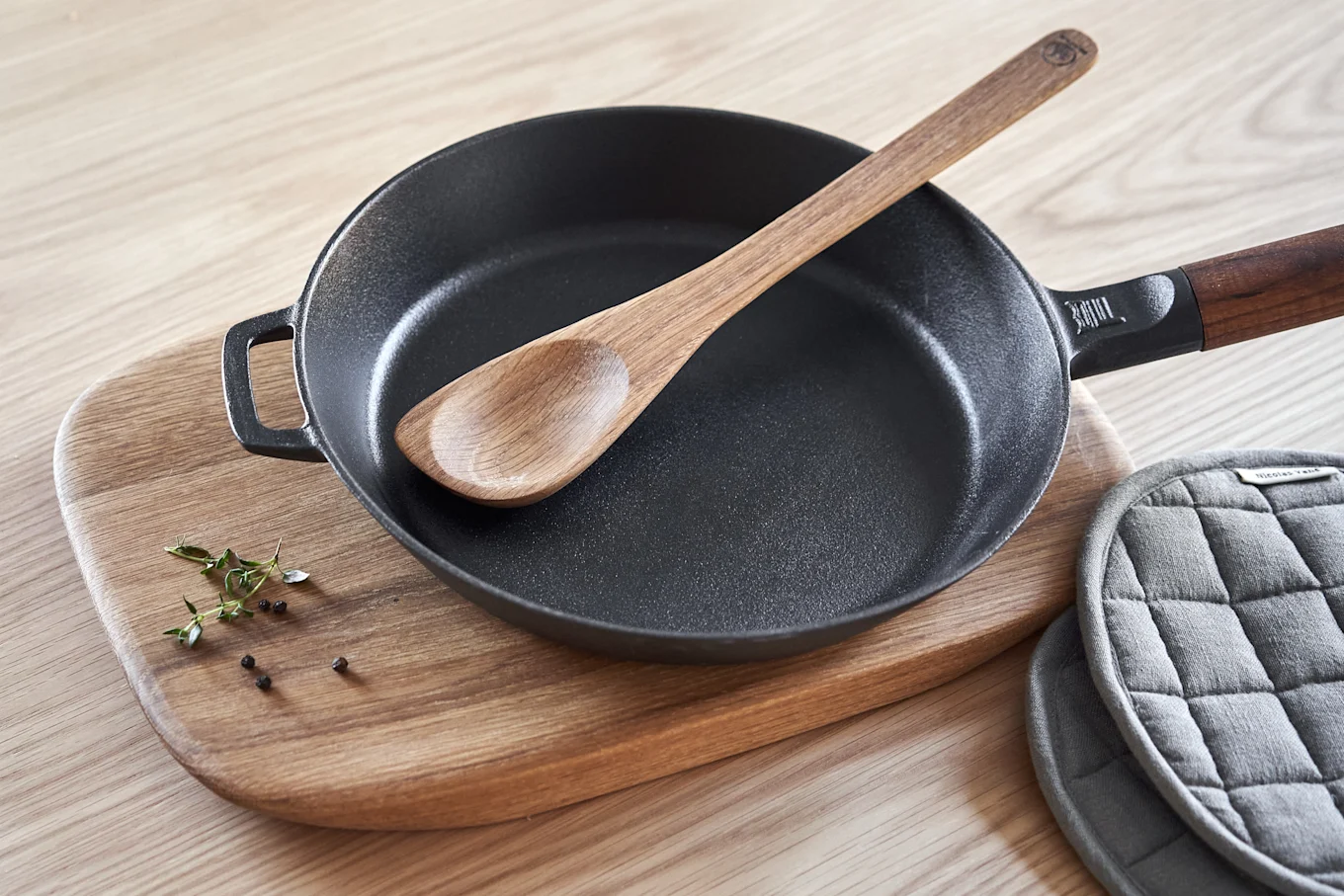 A black cast iron frying pan with a wooden spoon inside, on a wooden cutting board with herbs and peppercorns, next to grey pot holders.