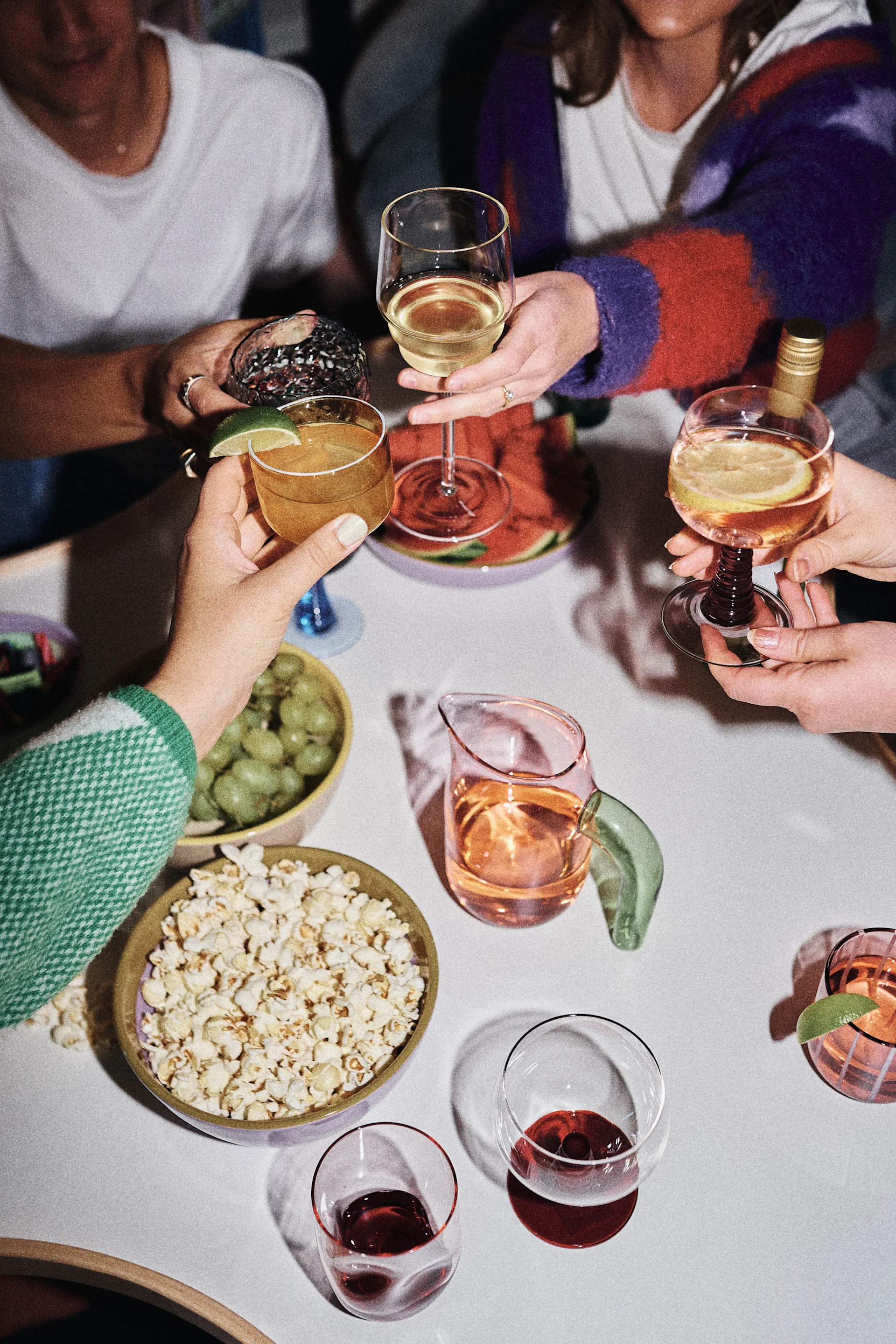 People clinking glasses with various drinks, surrounded by popcorn, grapes, and watermelon on a table.