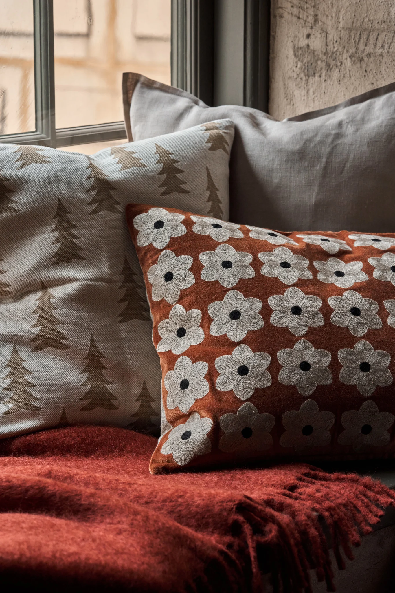 Pillows on a window seat: white with trees, orange with white flowers, and plain gray, on a fuzzy red blanket.