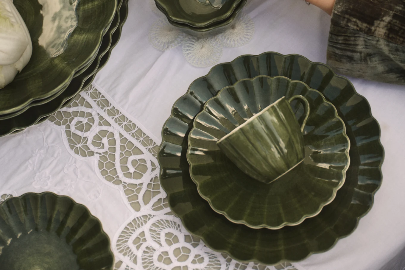 Dark green scalloped ceramic plates and cup on a white lace tablecloth with a white vegetable.