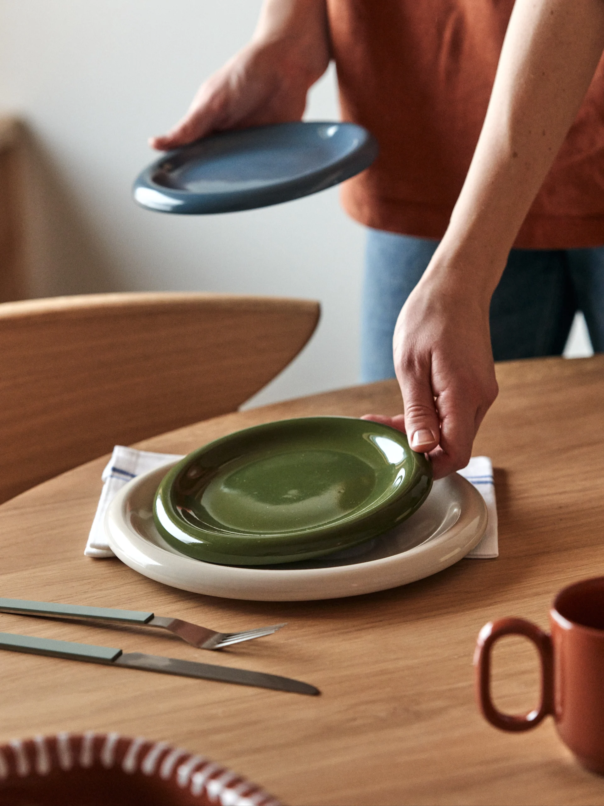 Hands placing a green plate on a beige plate at a wooden table, with another blue plate, cutlery, and mugs nearby.