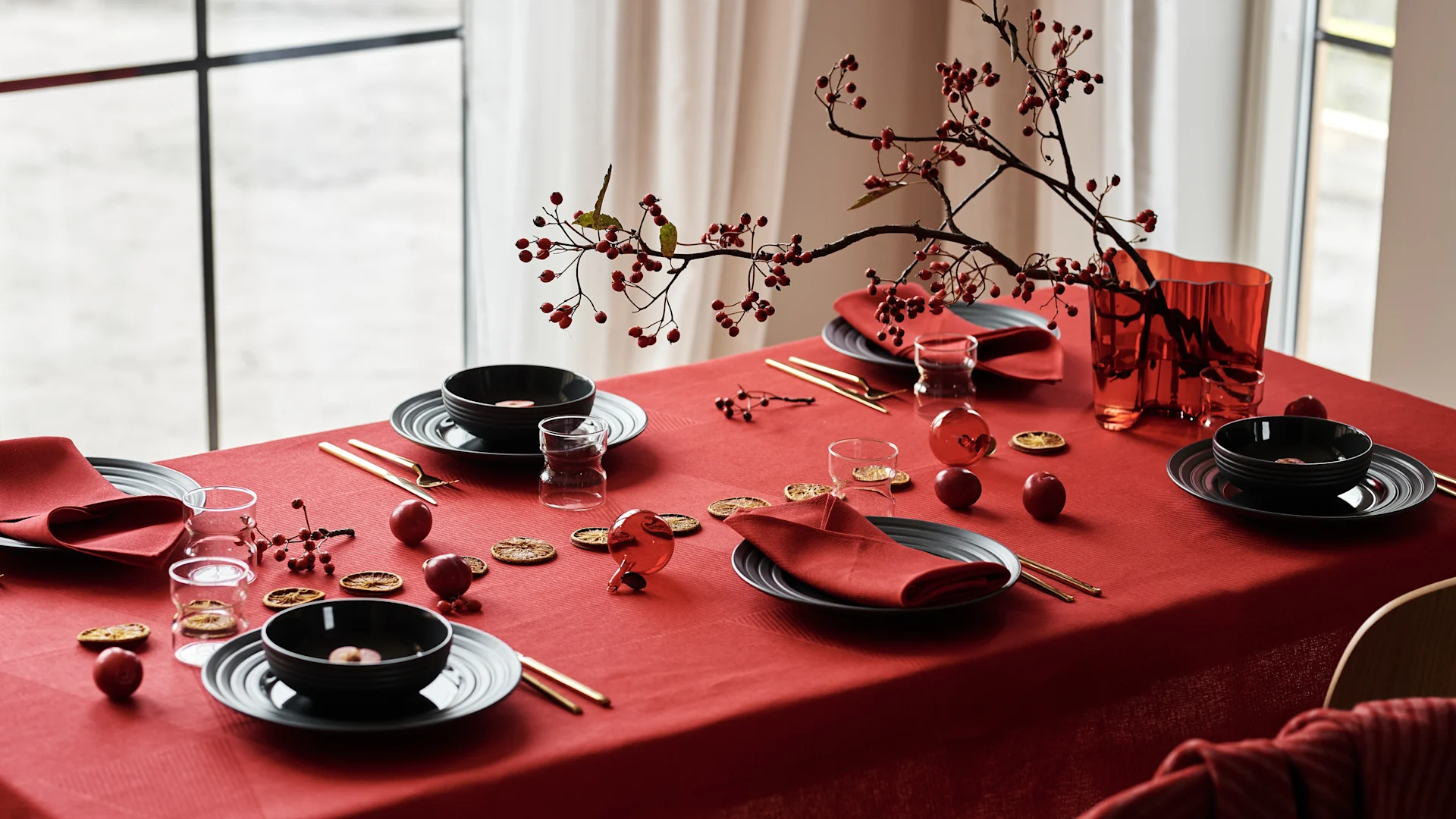 Festive table set with red tablecloth, black dishes, red napkins, gold cutlery, red berries, and dried orange slices.