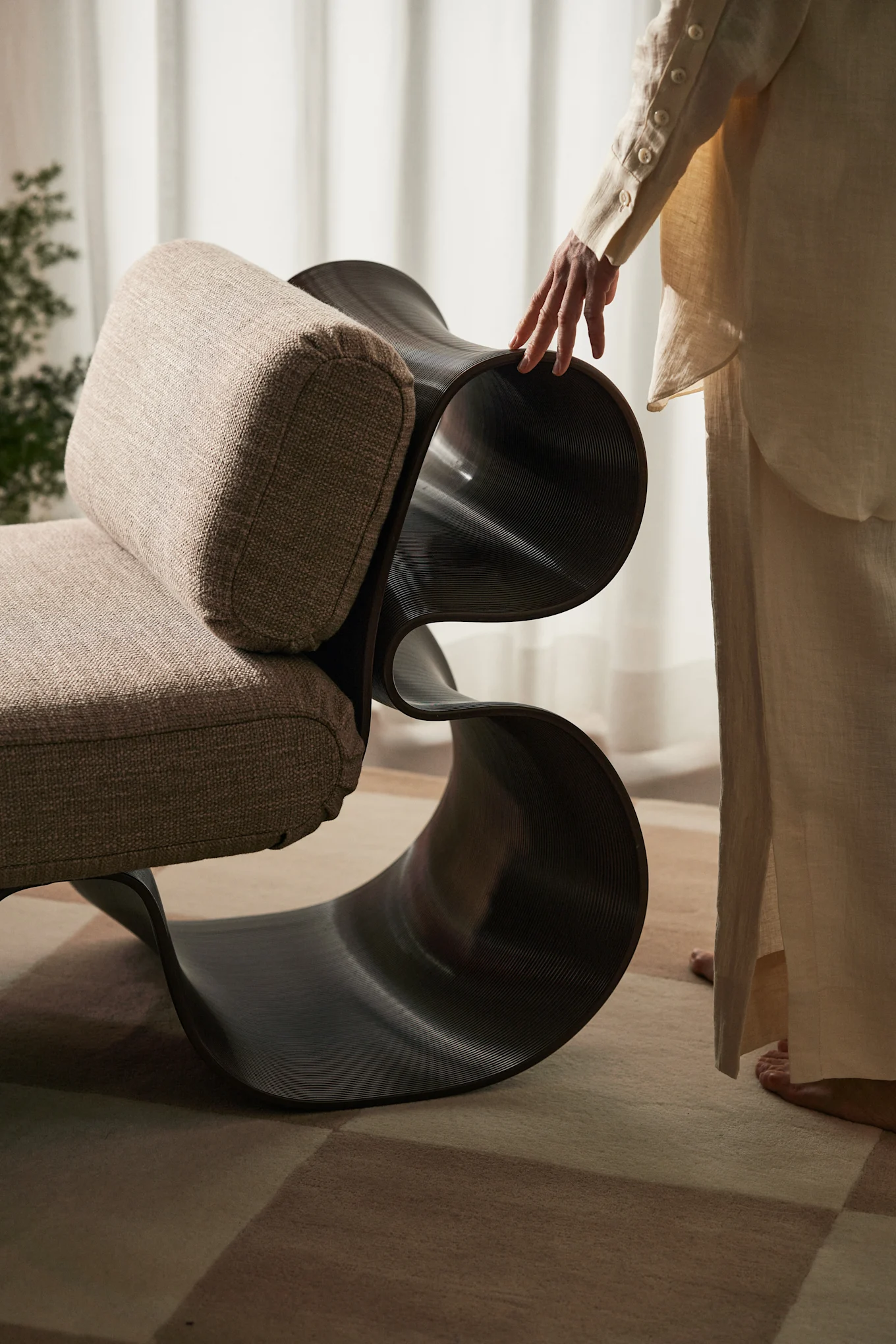 Person's hand touching a modern chair with a beige upholstered seat and dark, wavy, sculptural frame.