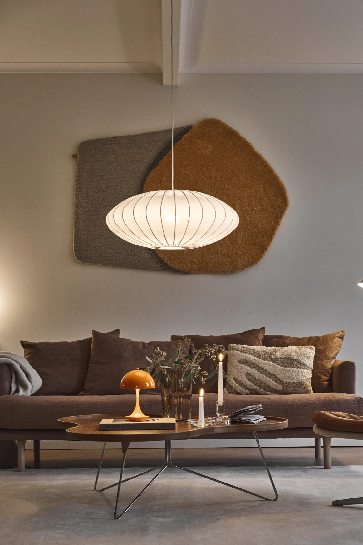 A cozy living room with a brown sofa, a dark wood coffee table with an orange mushroom lamp and candles, a white pendant light, and a brown leather lounge chair.