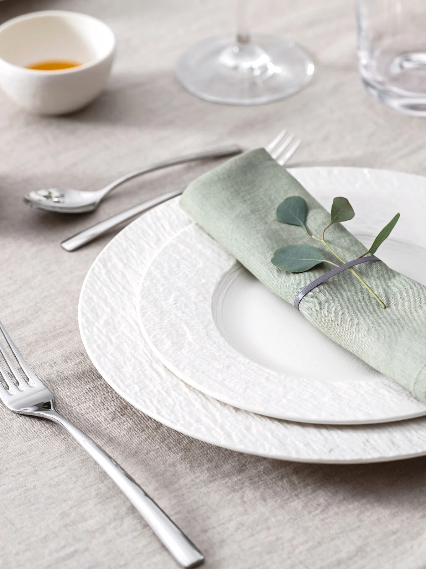 Elegant table setting with light green linen napkin, eucalyptus leaves, white textured plates, and silverware on a beige tablecloth.