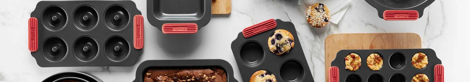 Assortment of dark gray baking pans with red handles on marble, including donut, loaf, and muffin pans with baked goods.