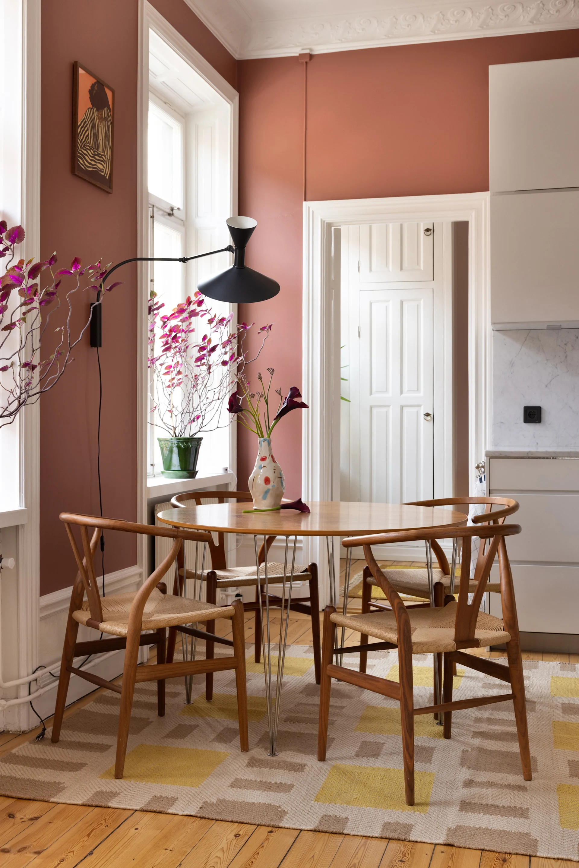 Dining area with a round wooden table, four wishbone chairs, a black wall lamp, a vase of calla lilies, and a framed art print on a rose wall.