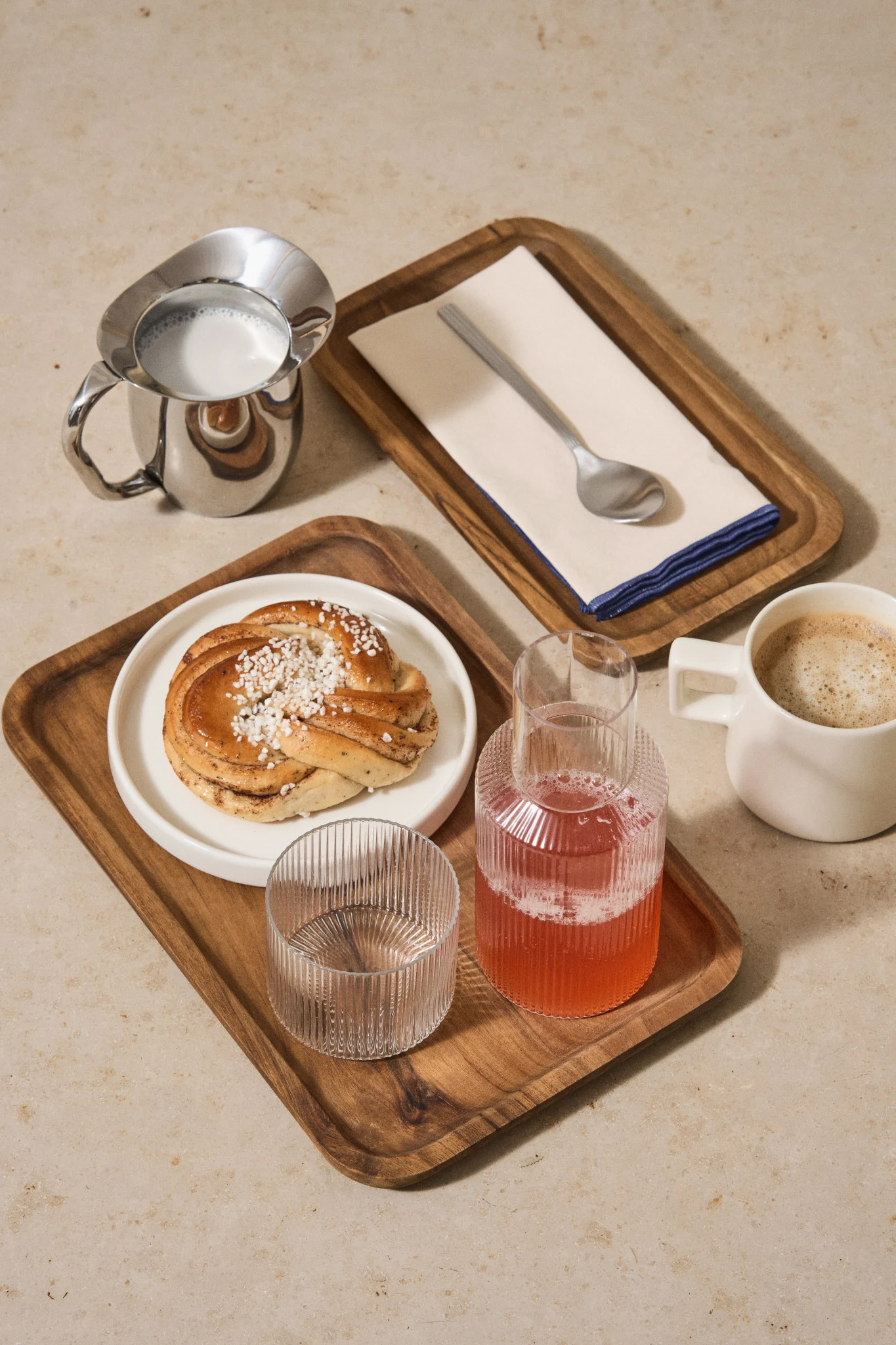 A cinnamon bun with pearl sugar, coffee, milk, a pink drink, and an empty glass on wooden trays.