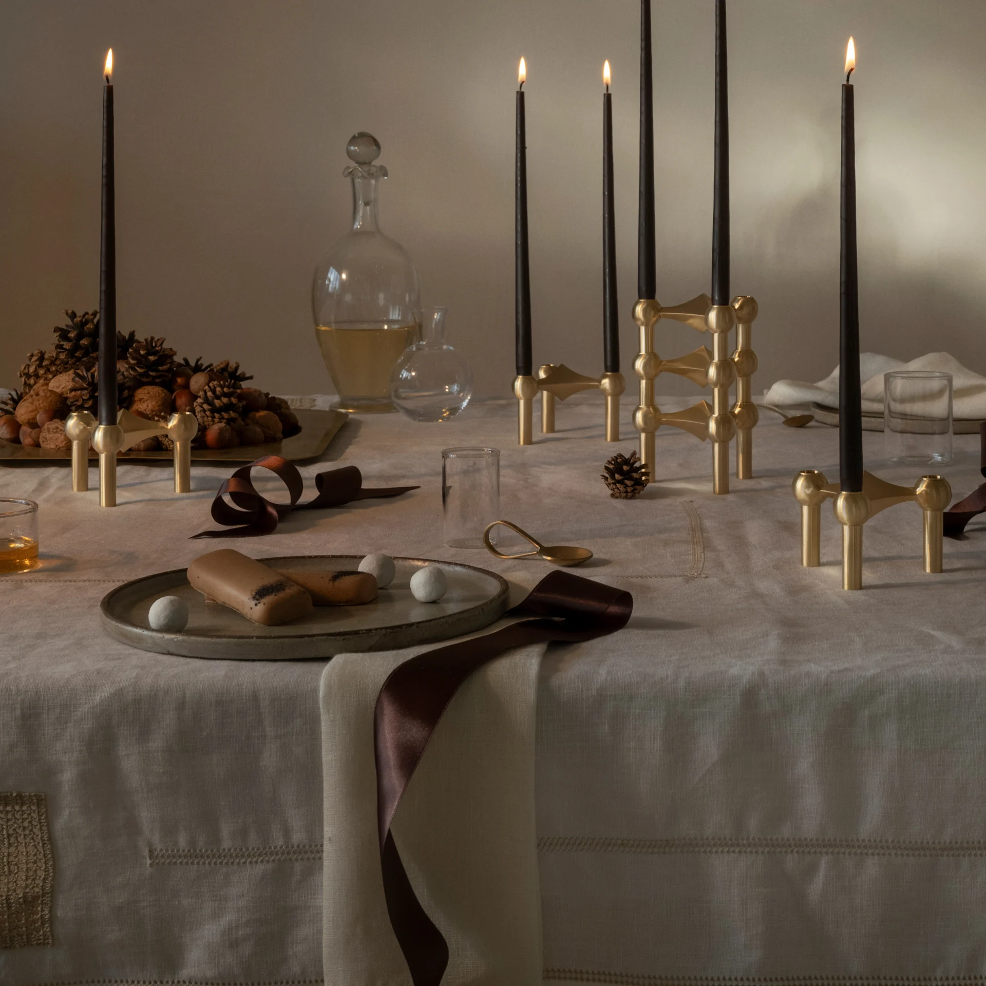 A holiday table set with lit black candles in brass modular holders, panettone, nuts, pinecones, and desserts on a linen tablecloth.