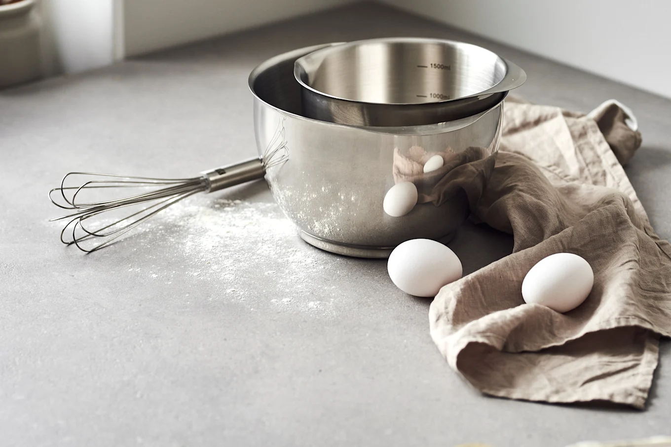 Stainless steel mixing bowls, a whisk, and three white eggs on a kitchen counter with spilled flour, ready for baking.