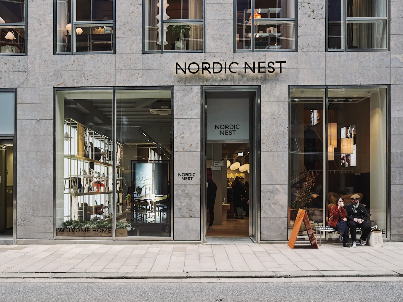 Modern Nordic Nest storefront with large windows displaying home furnishings, and two people sitting on a bench outside.
