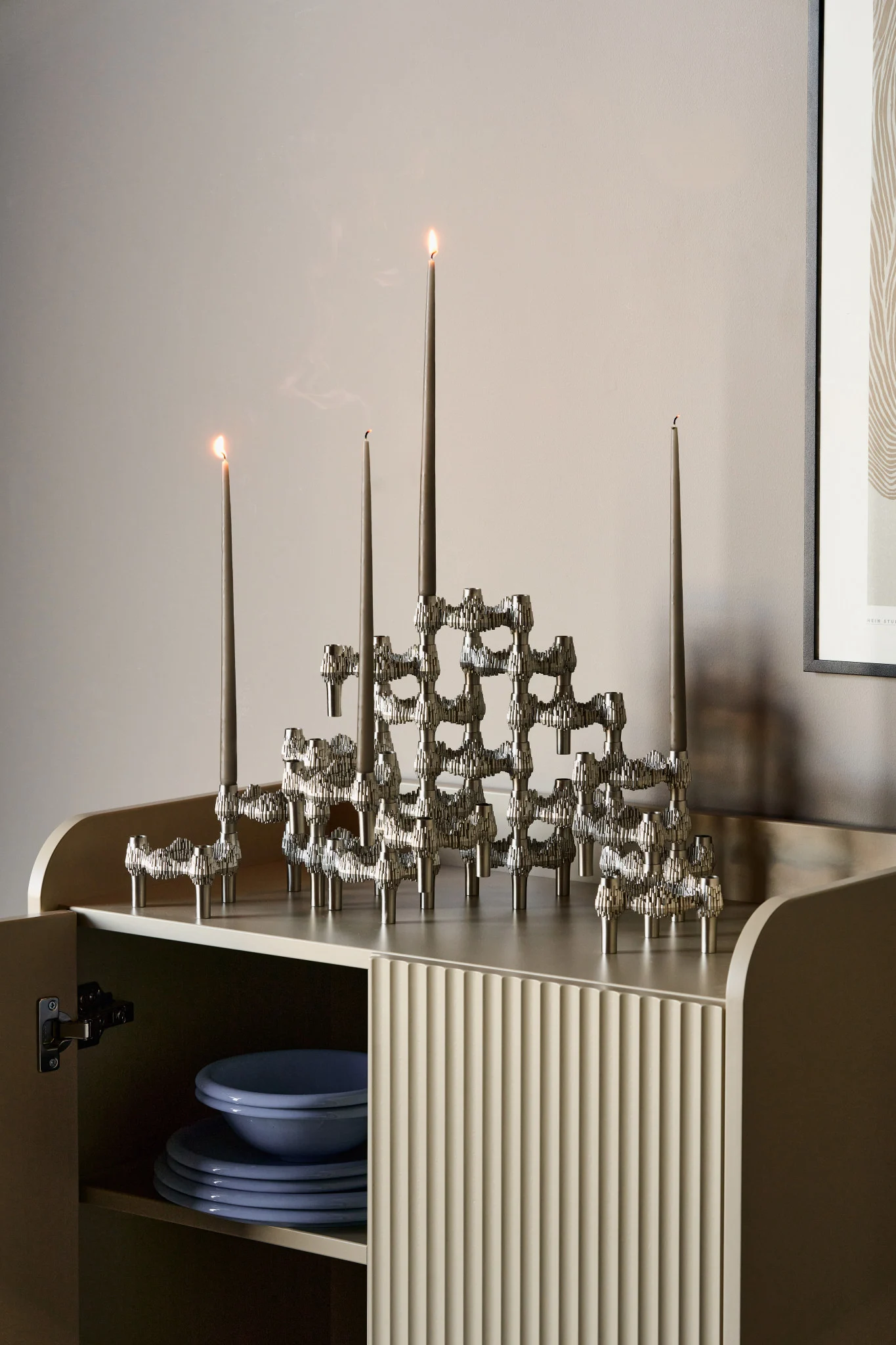 A modernist silver candle holder with tall grey taper candles, some lit, on a light fluted cabinet with blue dinnerware.