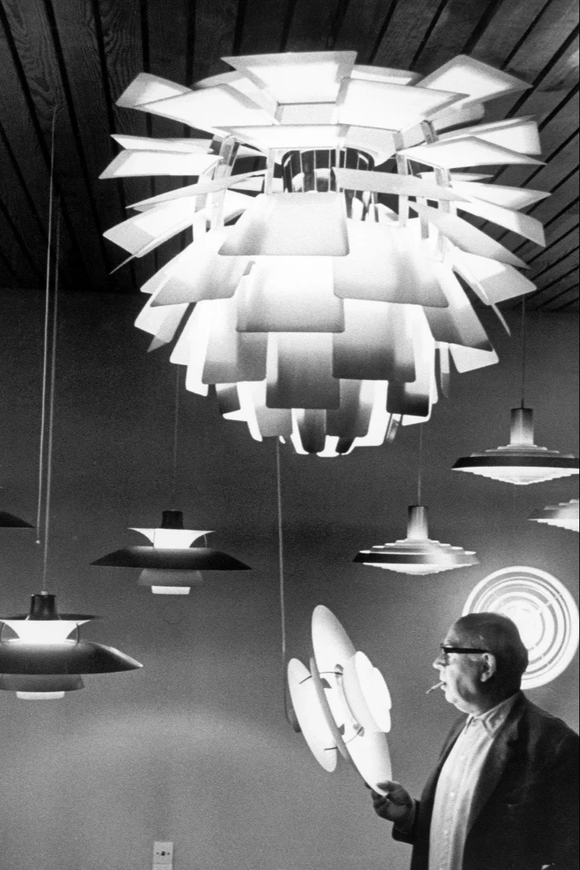 Man in glasses smoking, holding a disc-shaped lamp and looking at several unique layered pendant lights.