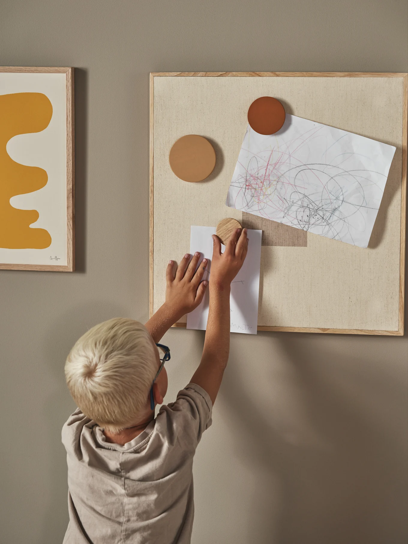 Child pinning a drawing to a fabric bulletin board with a round wooden magnet, beside an abstract art print.