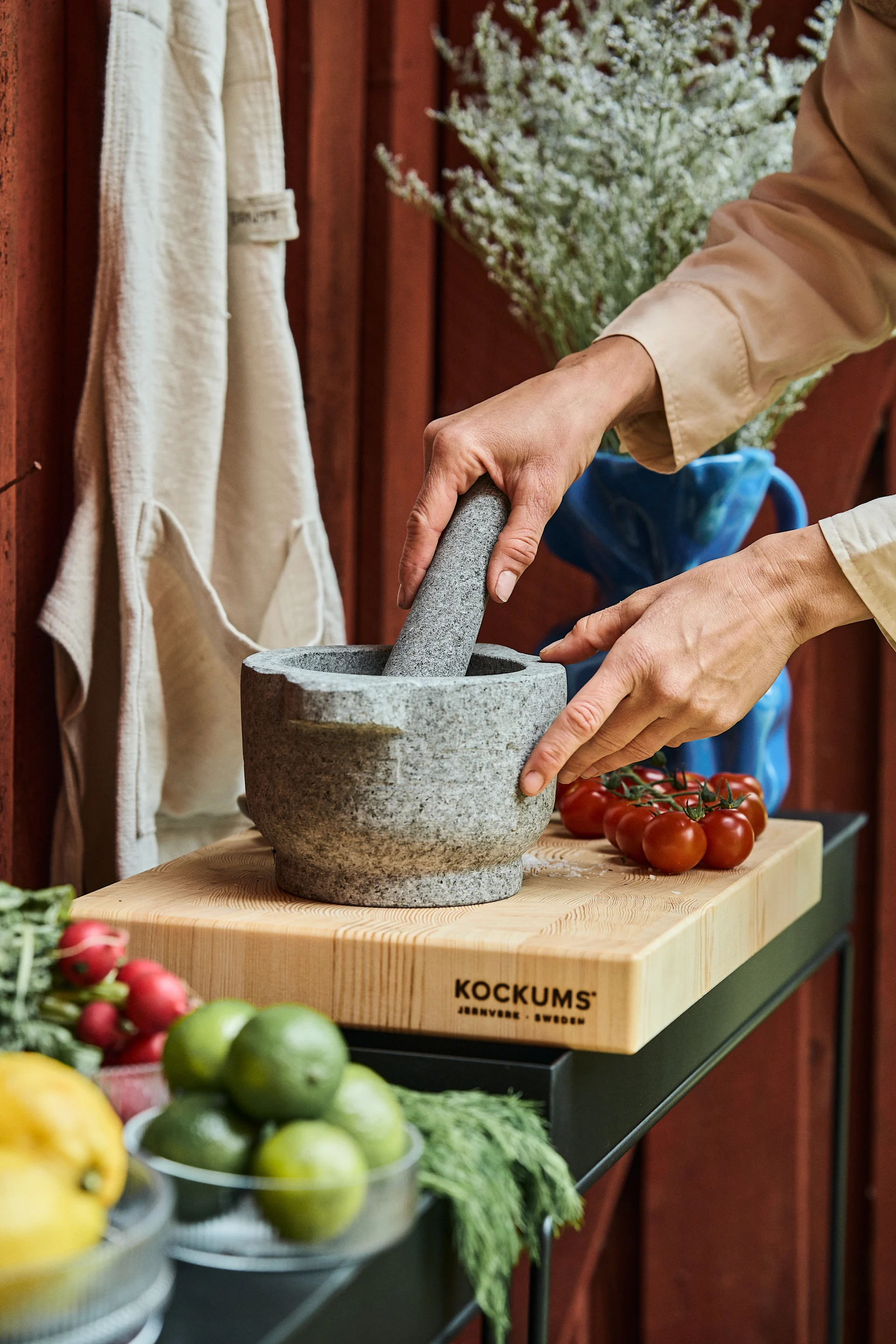Hands grinding spices in a grey stone mortar and pestle on a wooden cutting board with fresh vegetables.