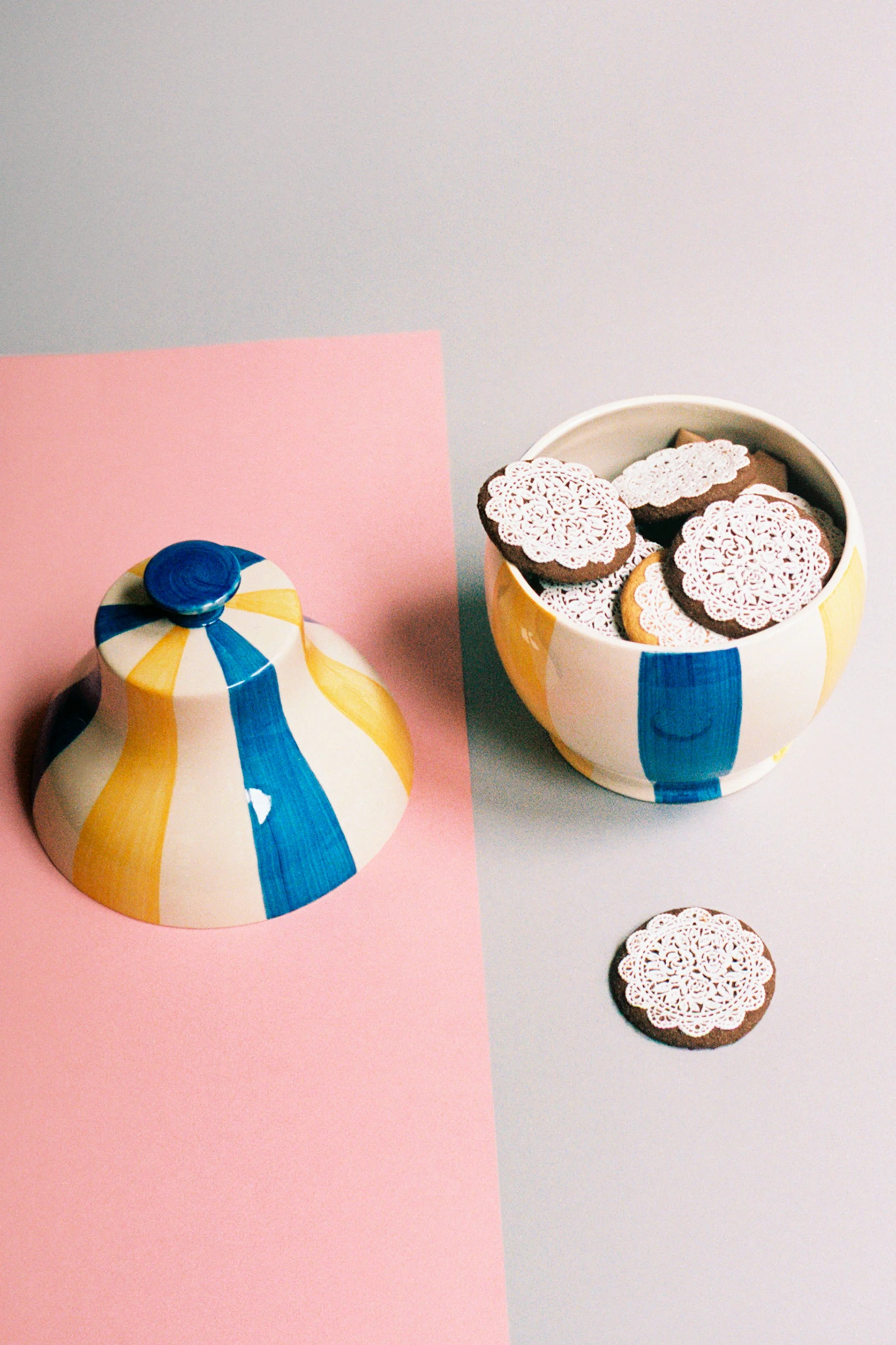 Striped blue and yellow ceramic cookie jar and bowl filled with chocolate cookies, each topped with intricate white lace-like icing.