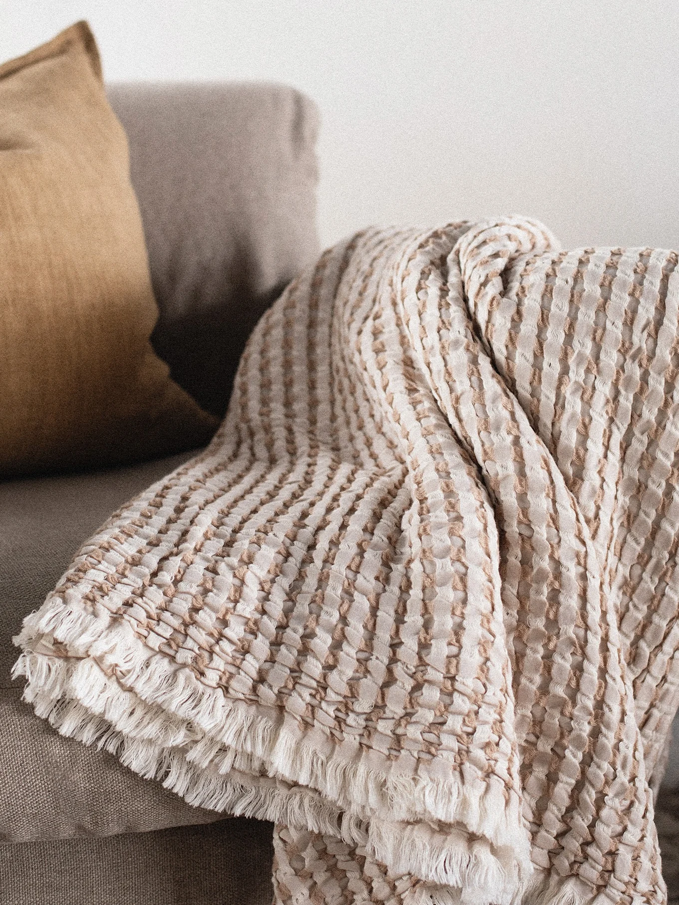 Textured light beige and brown striped throw blanket with fringe draped on a sofa next to a mustard pillow.