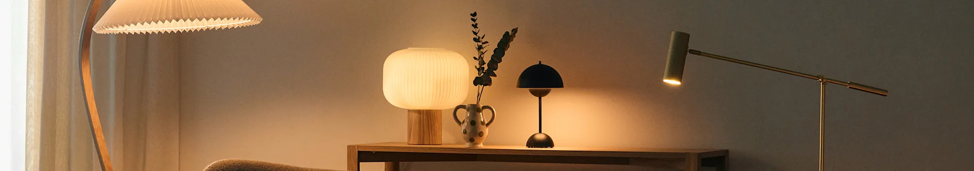 A cozy room with various lamps including a pleated floor lamp, a ribbed table lamp, a small black table lamp, and a brass floor lamp.
