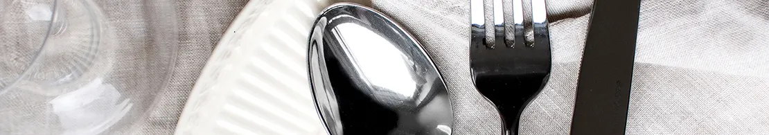 A close-up of a modern table setting with a clear glass, white ridged plate, silver spoon, black fork, and black knife on light fabric.