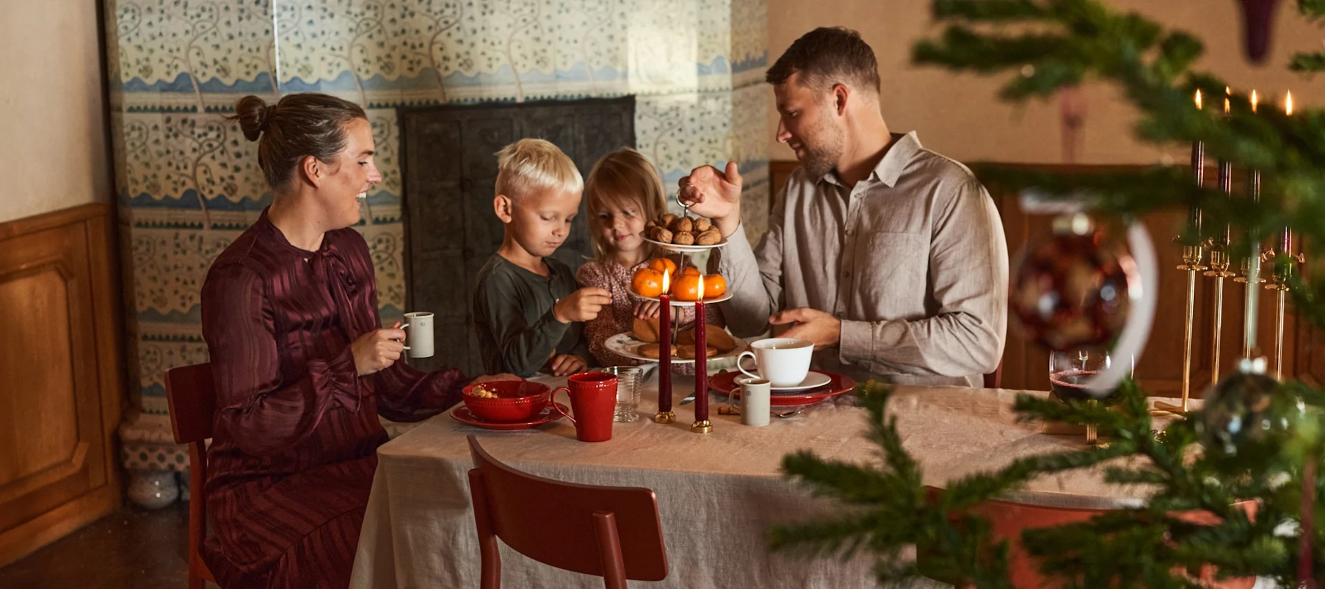 Family enjoying a festive meal with children, nuts, mandarins, and candles at a table with a Christmas tree.