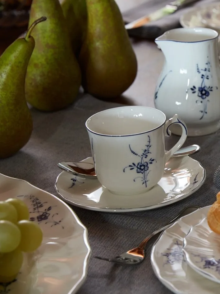 White porcelain teacup, saucer, and pitcher with blue floral pattern on a table with green pears and grapes.