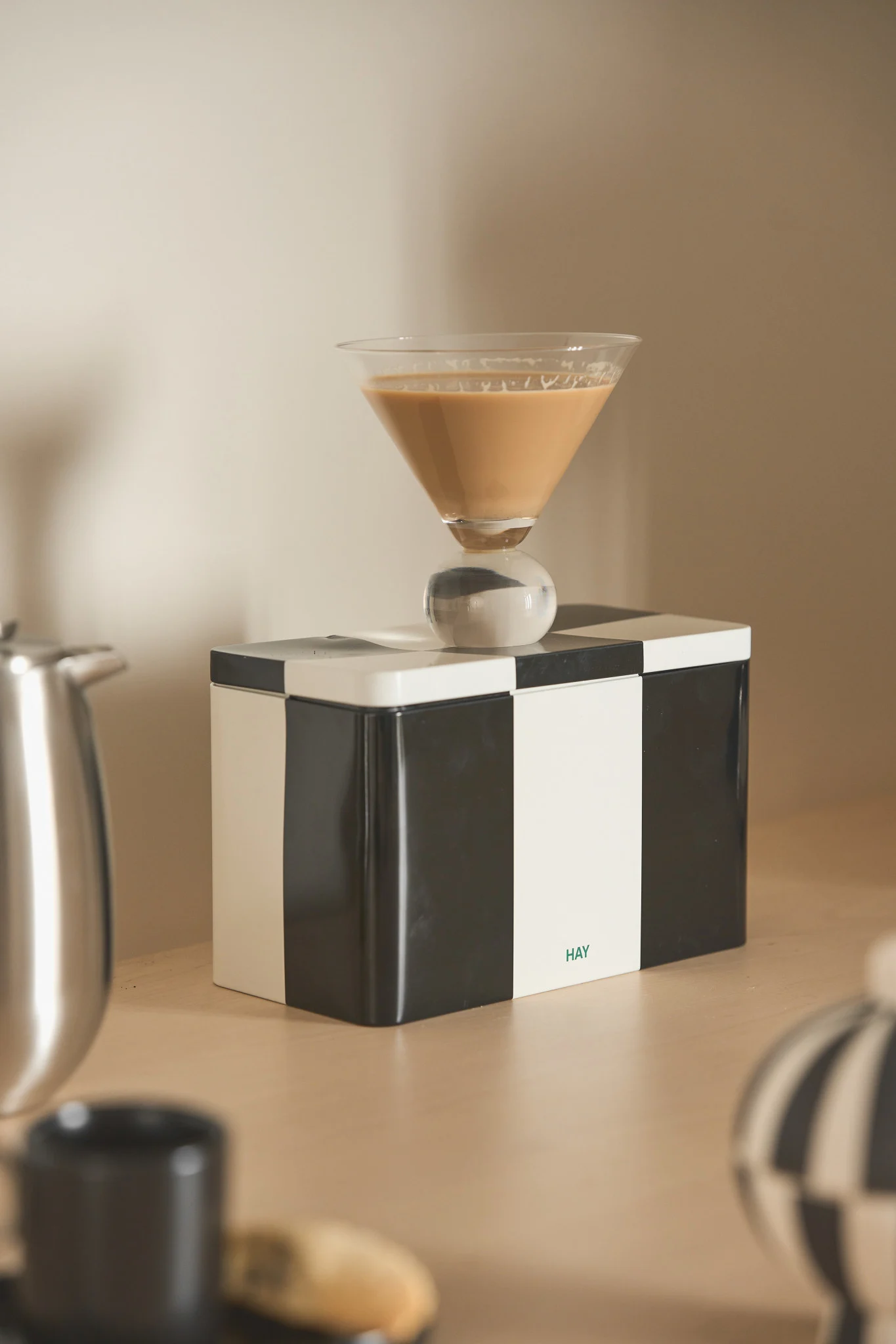 Martini glass with a light brown drink and spherical stem on a black and white checkered box with a 'HAY' logo.