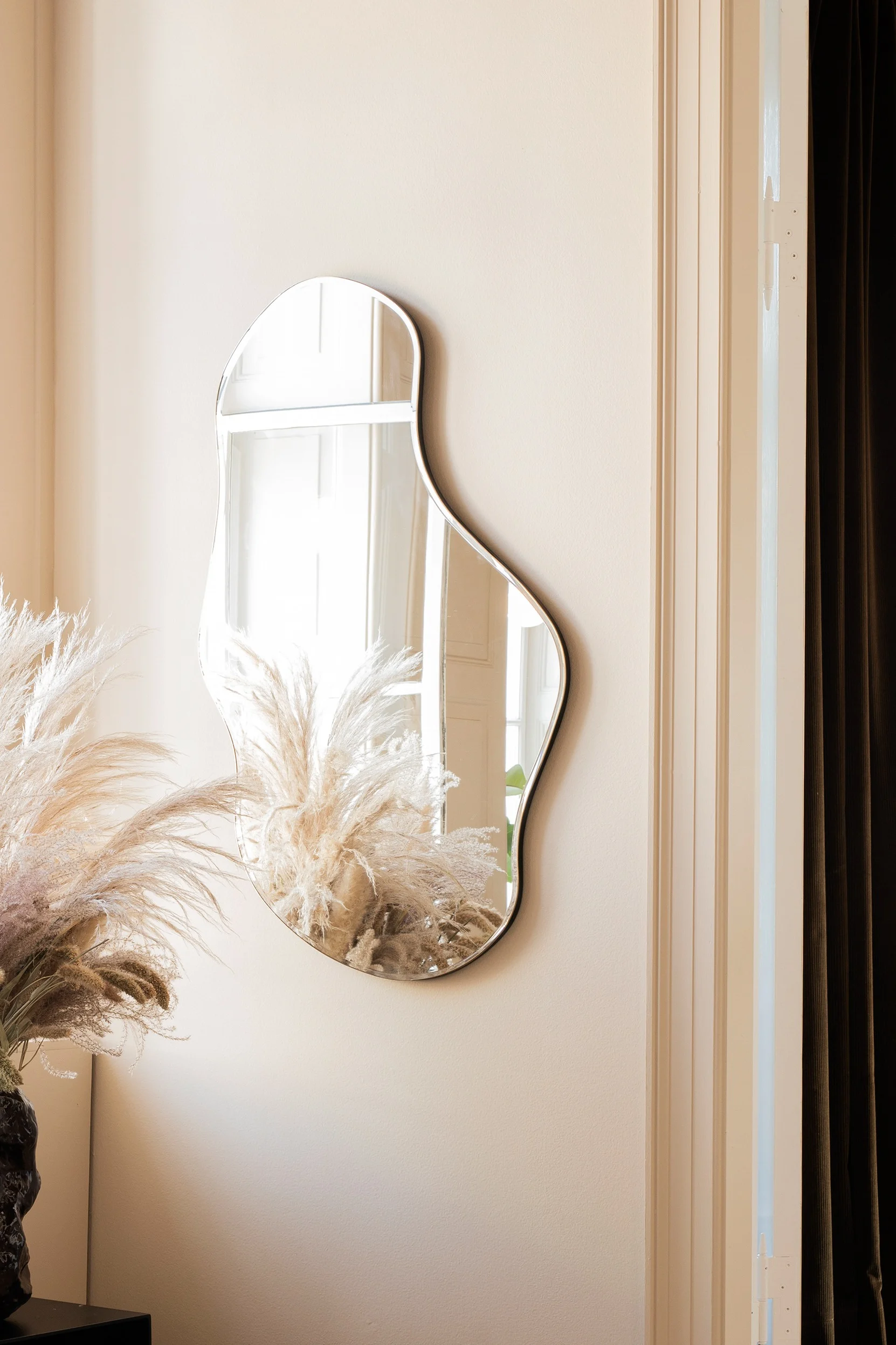 A room corner with a large window, a black table with a vase of dried pampas grass, and a wavy mirror.