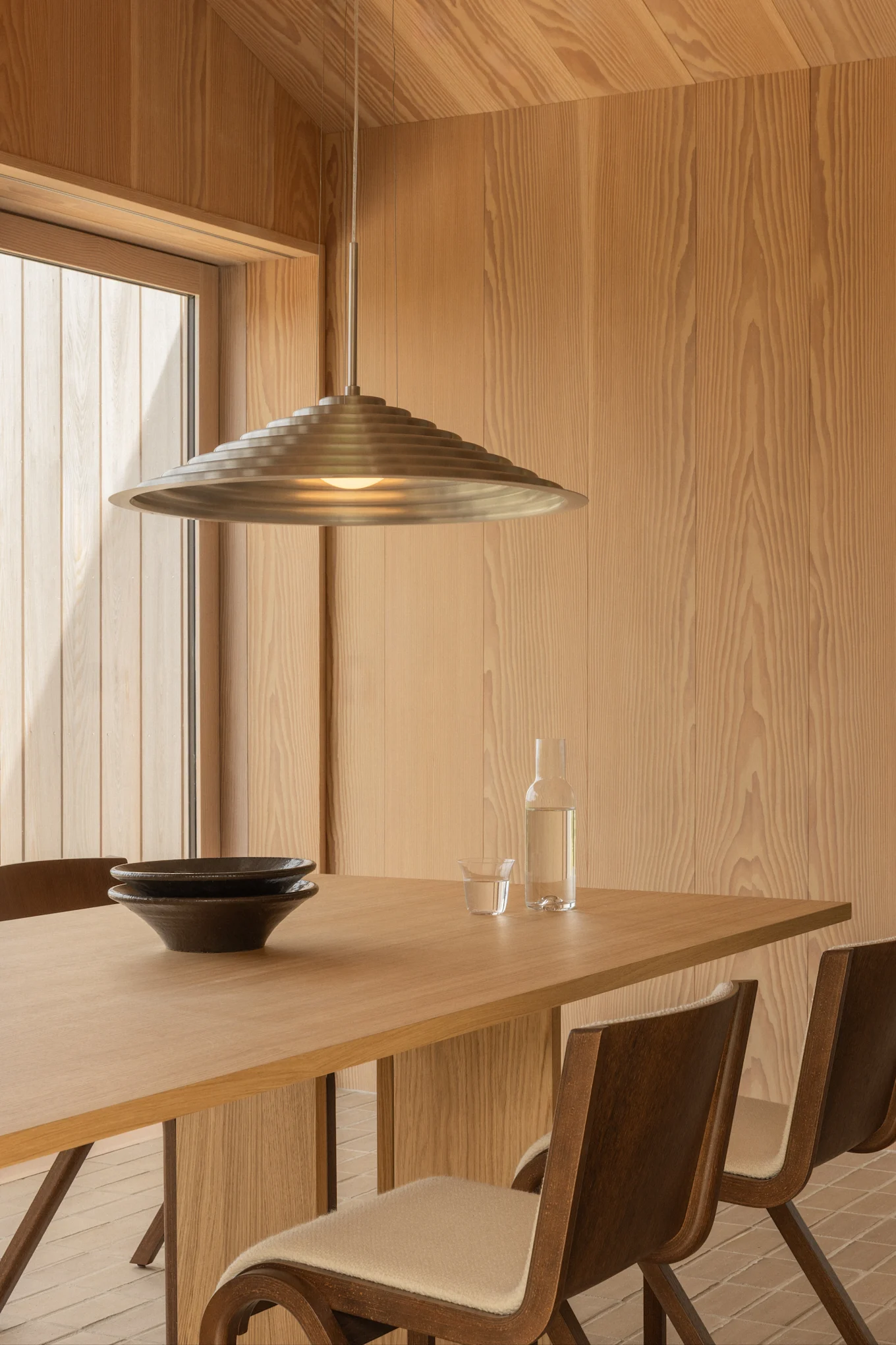 Modern dining room features light wood walls, a wooden table with two dark bowls, a water bottle, and a tiered metal pendant lamp.