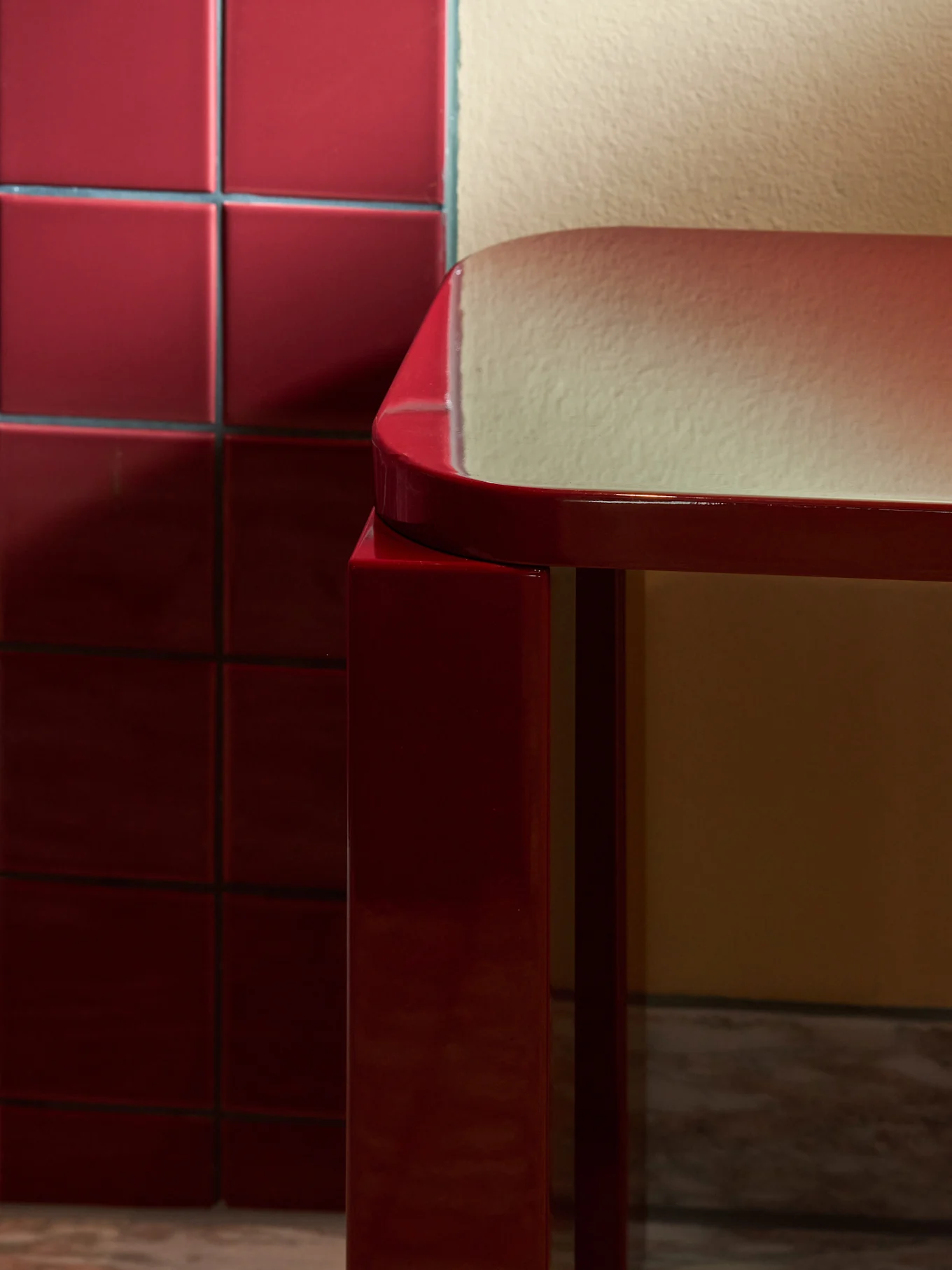 Sleek, shiny red rectangular stool with rounded edges on a marble floor, next to a red tiled wall.
