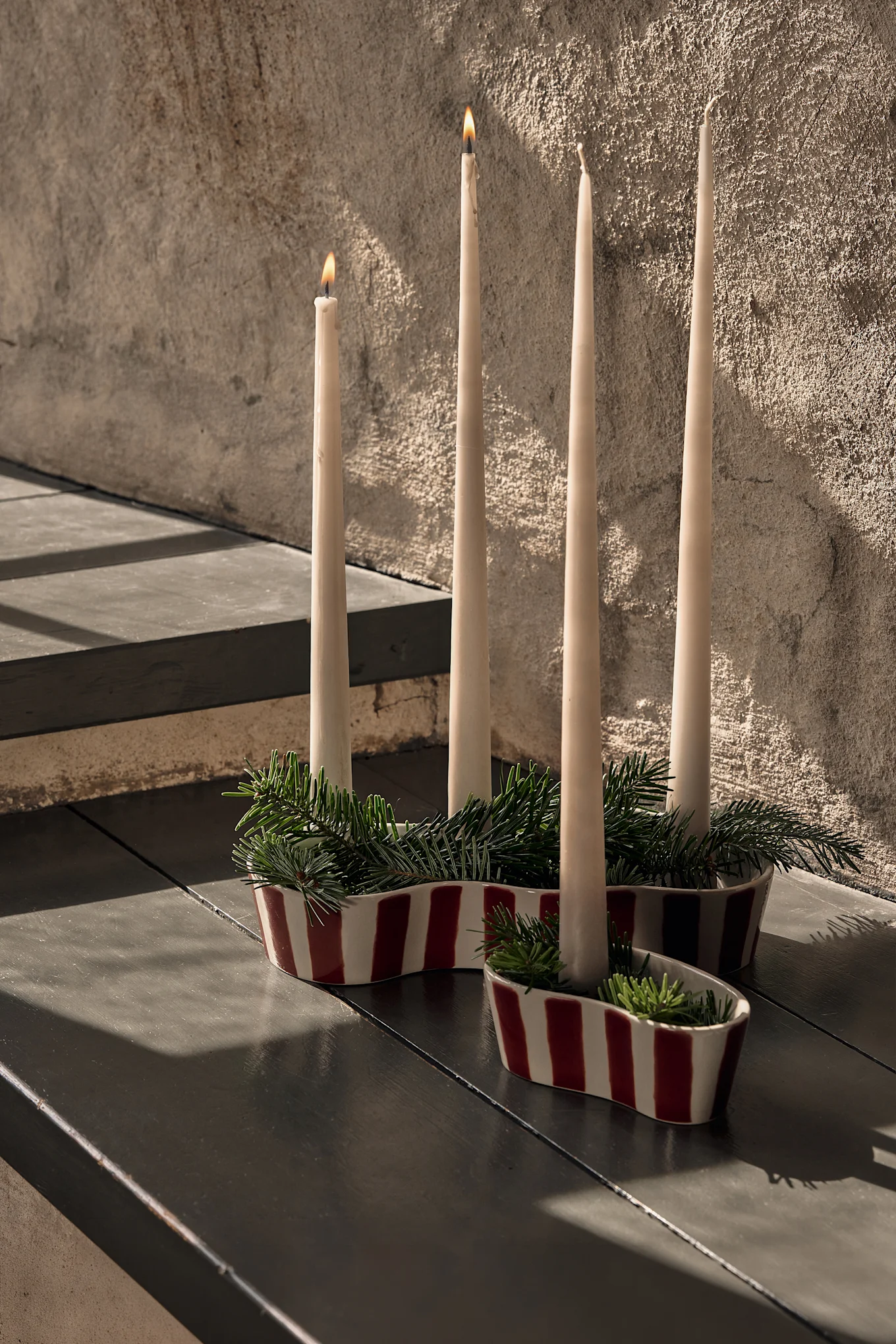 Four cream candles in red and white striped ceramic holders with pine branches, two candles lit, on a dark table.