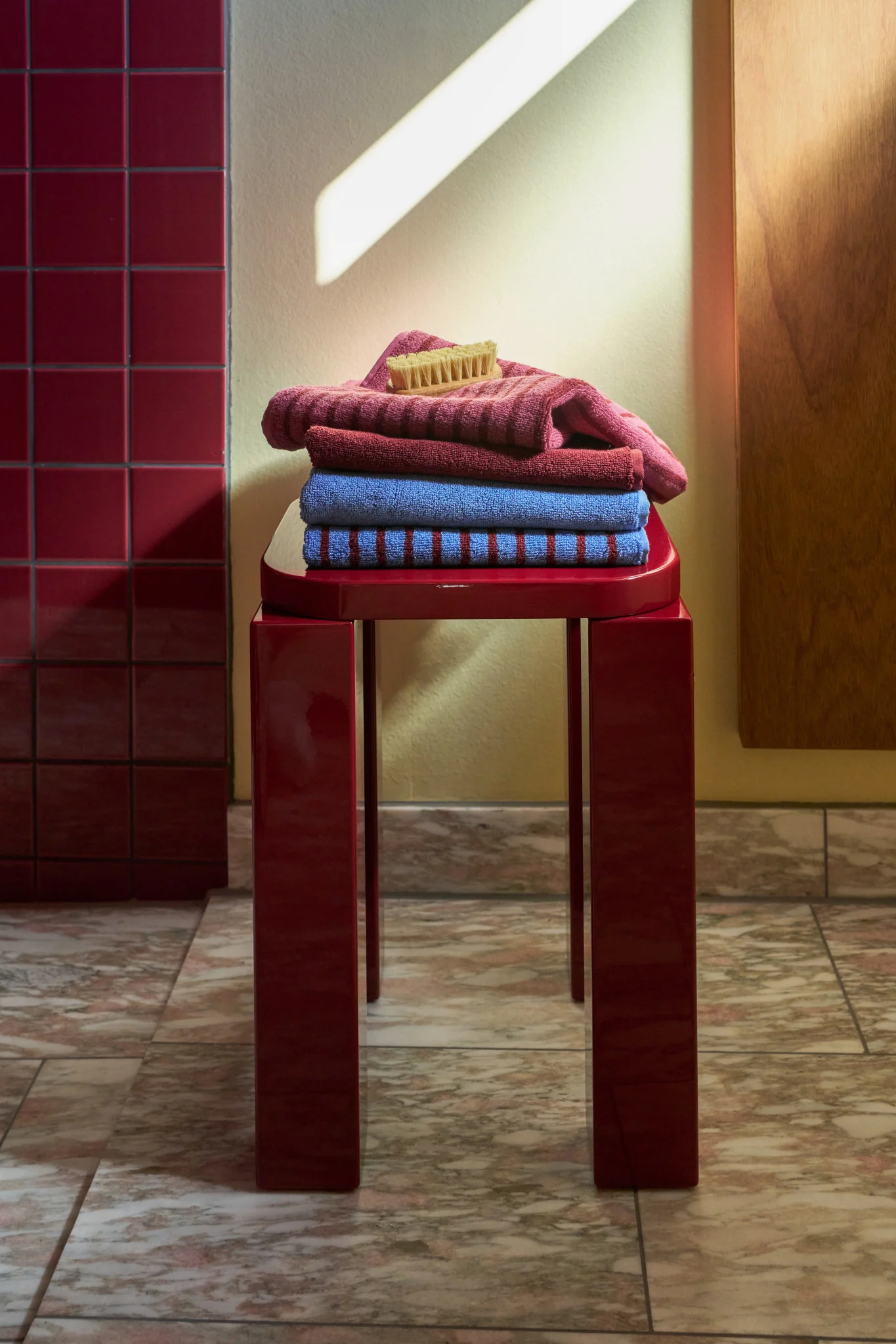 A stack of pink, red, and blue towels with a brush on top, on a red stool in a tiled room.