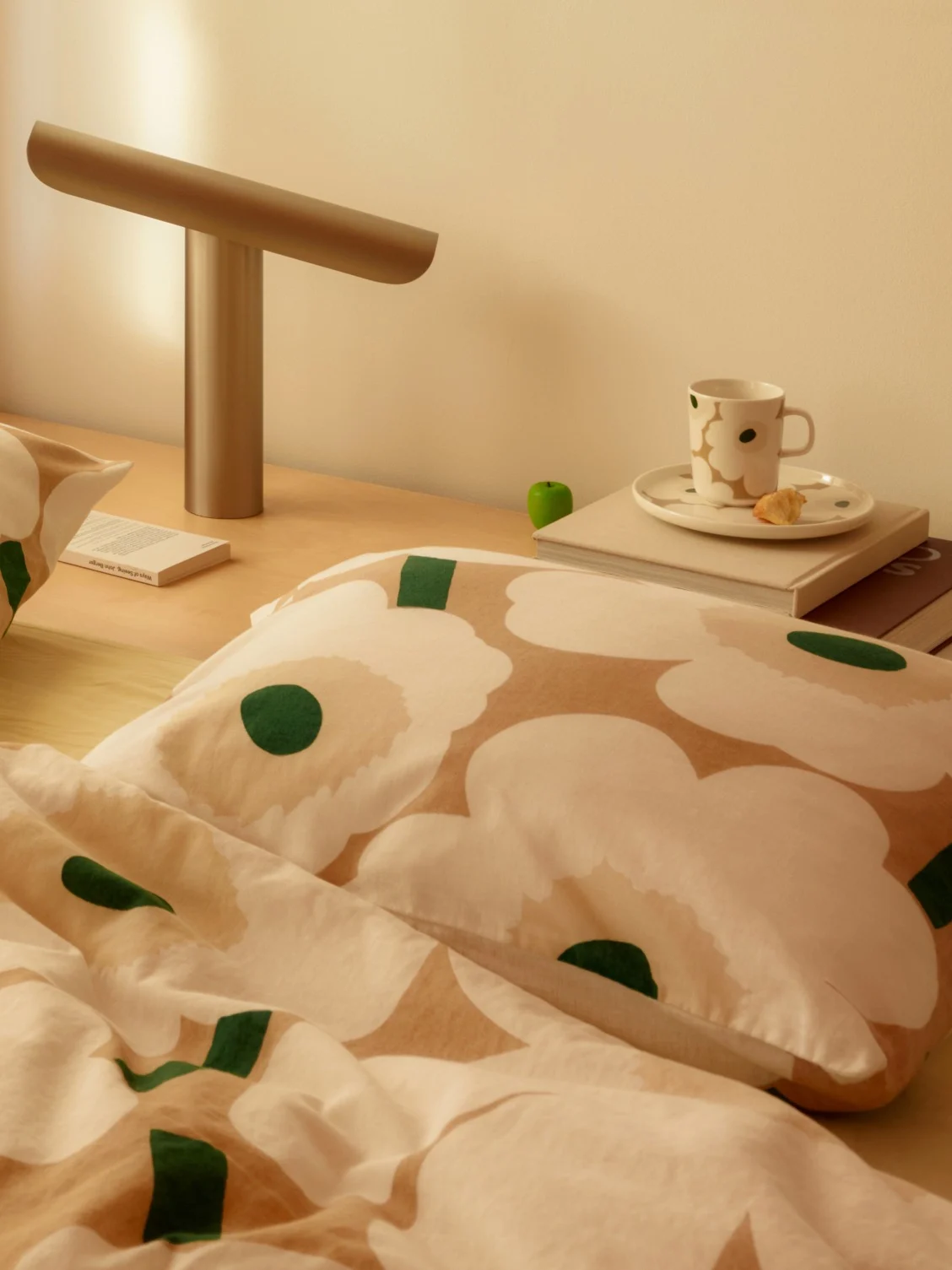 Bedside scene with white and beige floral bedding, a modern lamp, a cup, and a book on a wooden nightstand.
