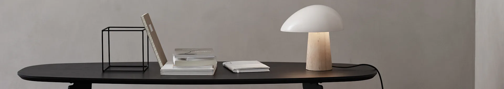 A minimalist desk with a black table, black metal stand, stacked books, and a lamp with a wood base and white mushroom shade.