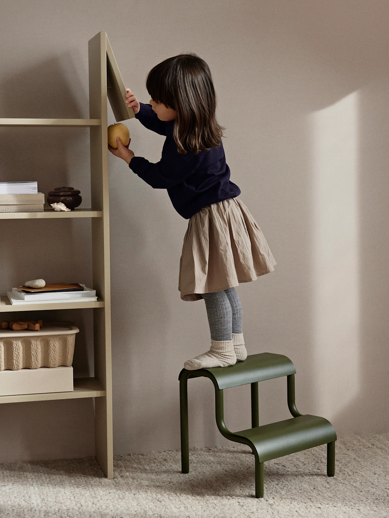A young child standing on a green two-step stool, reaching for a yellow apple on a light beige shelf.