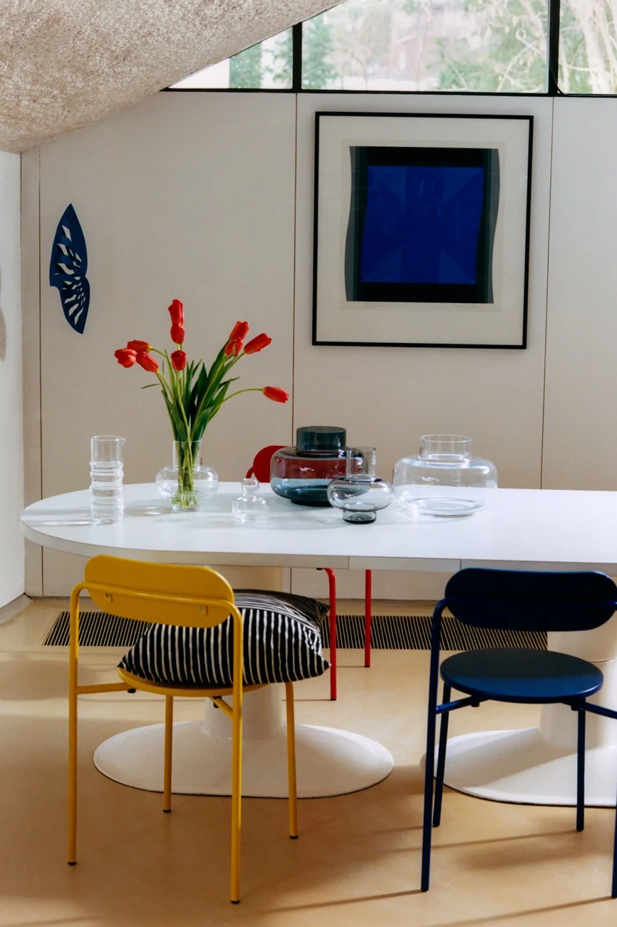 Modern dining room with white oval table, red tulips, glass vases, yellow and blue chairs, and abstract blue art.
