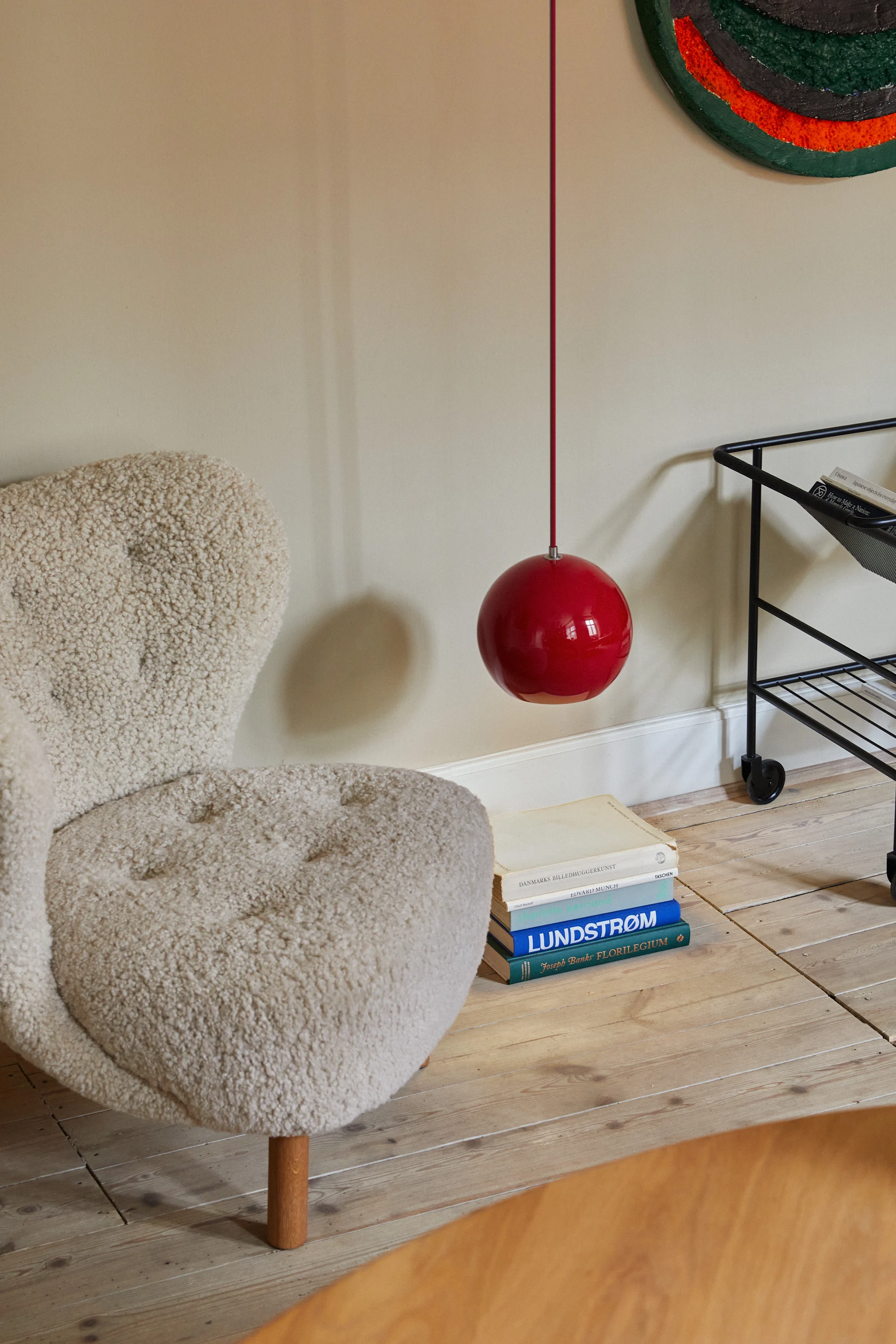 A fluffy white armchair, red spherical pendant lamp, and stack of books on a wooden floor.