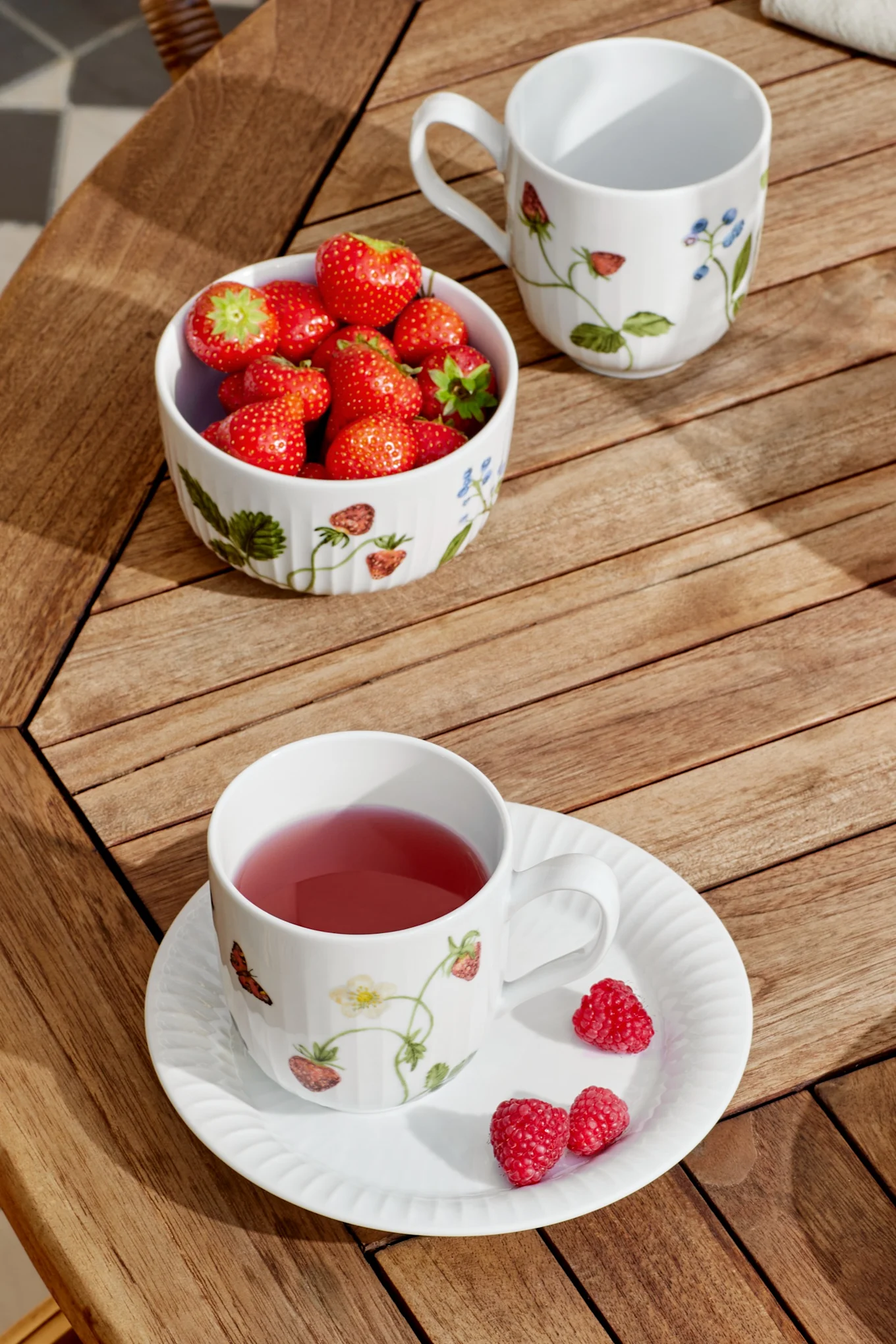A white mug with pink tea and a saucer with raspberries, a bowl of strawberries, and an empty mug sit on a wooden slatted table in sunlight.