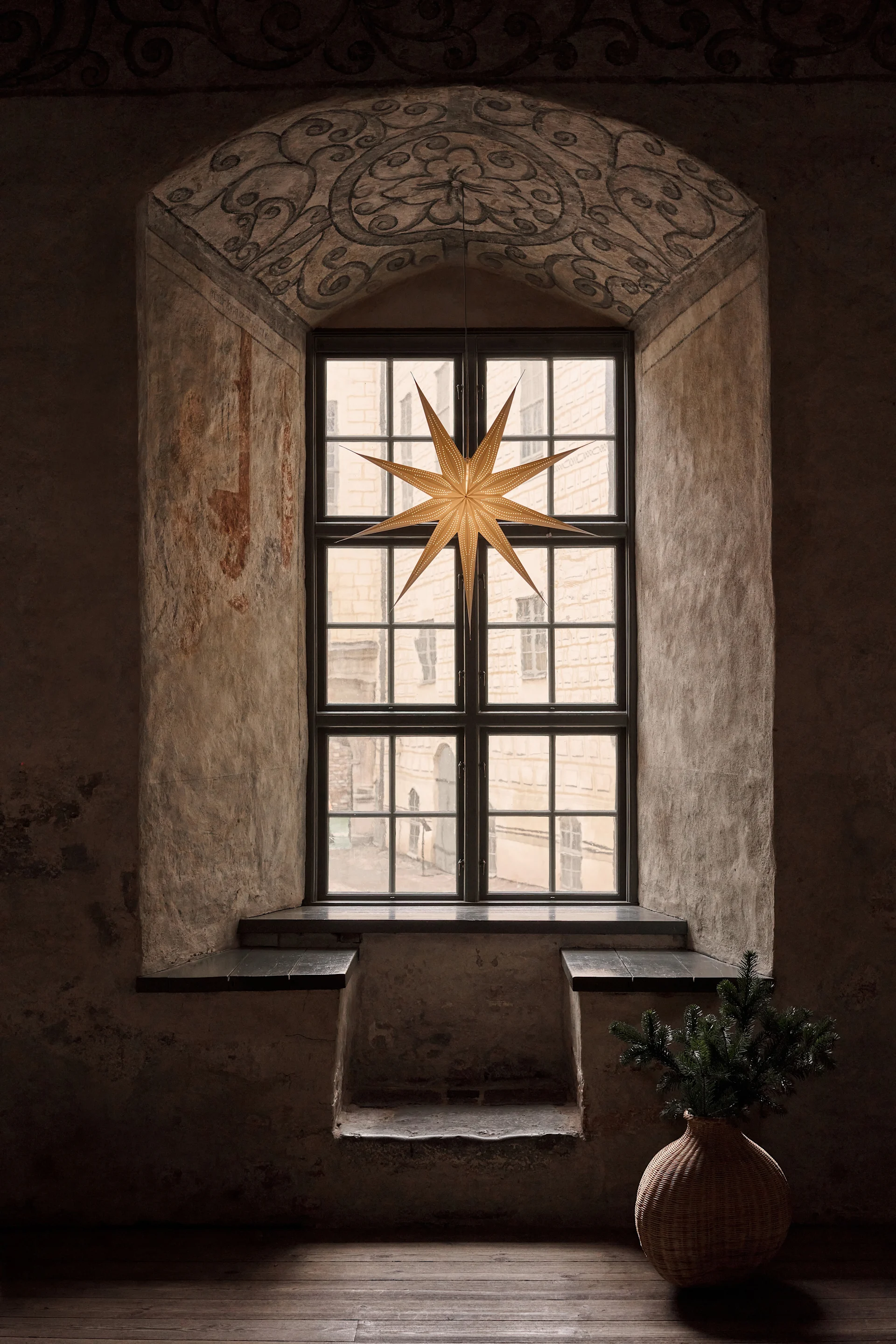 An old arched window with a large paper star hanging in it. A wicker vase with pine branches sits on the floor to the right.