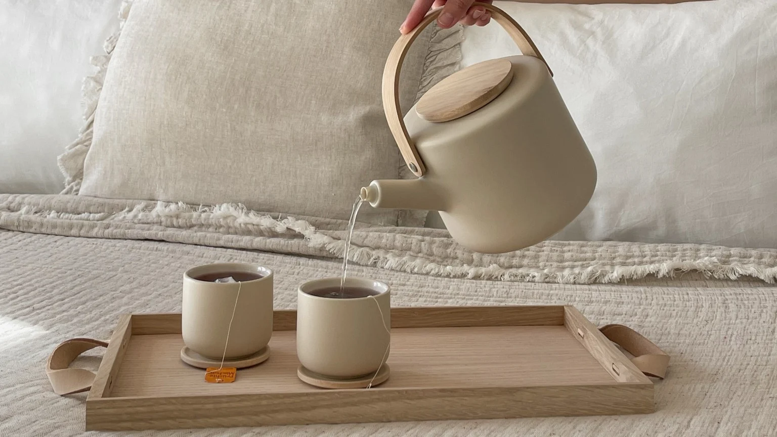 Hand pouring tea from a beige teapot with a wooden handle into a matching cup on a wooden tray in bed.