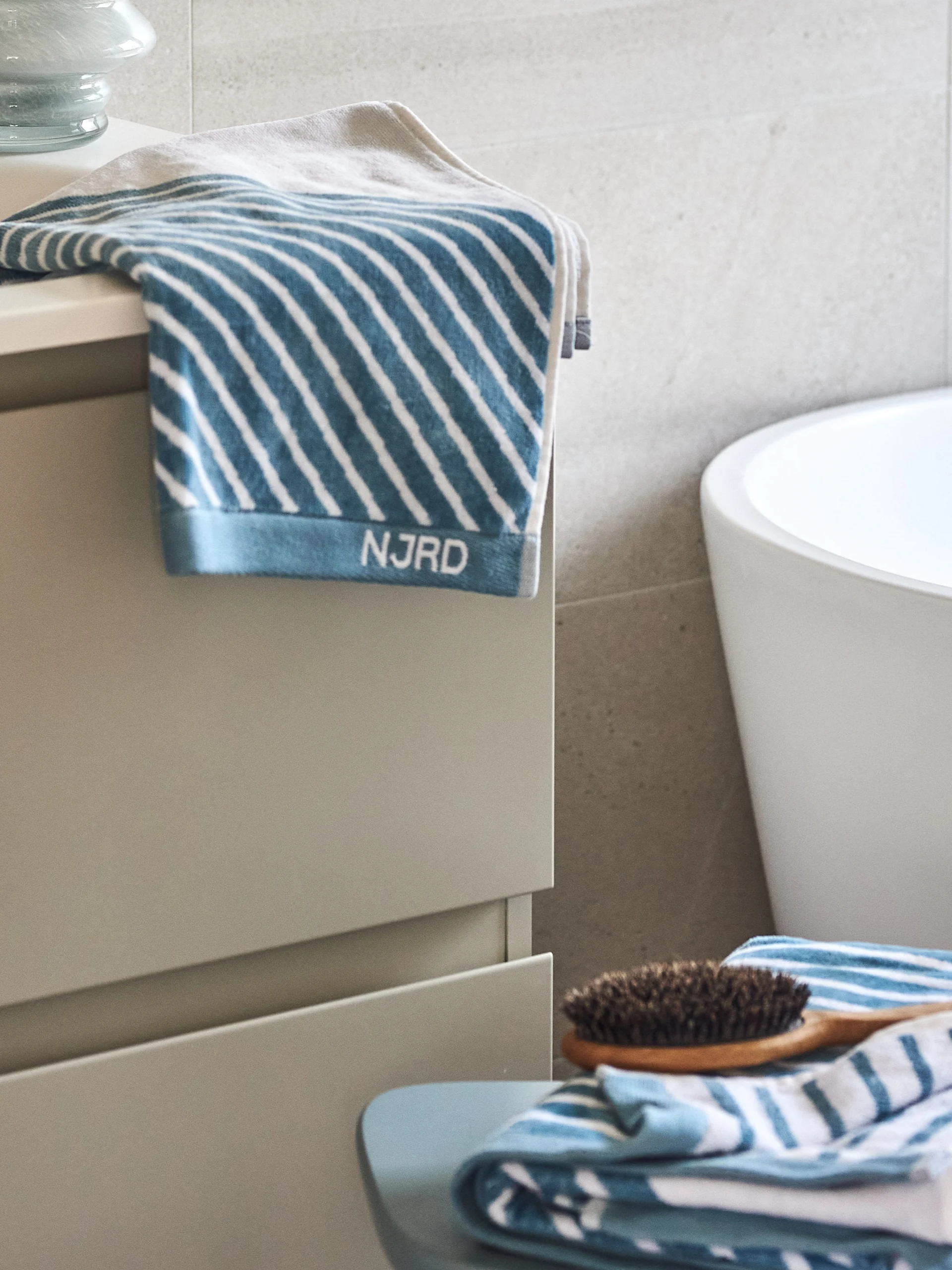 Blue and white striped towels and a wooden hairbrush in a bathroom with a white tub and grey cabinet.